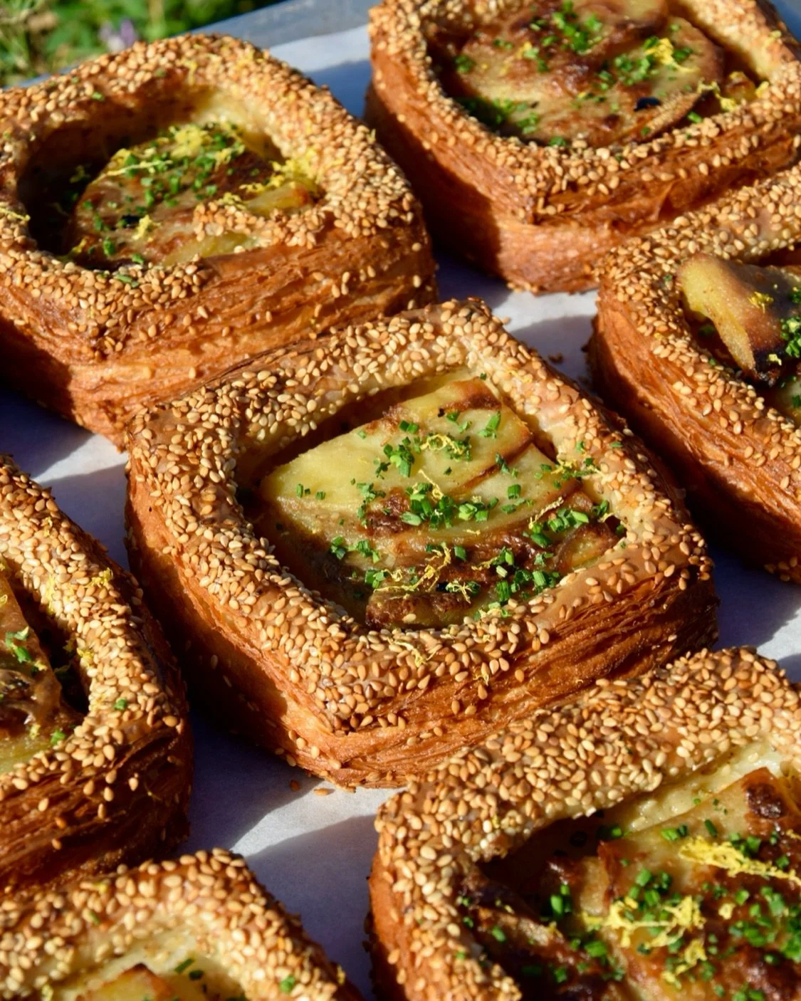 Close-up of several square-shaped pastries with flaky layers, sesame seed crust, topped with chopped herbs and glaze.
