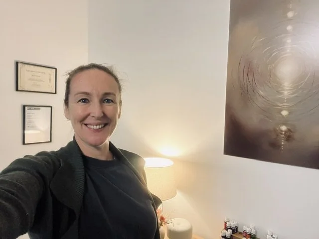A woman taking a selfie in a room with framed certificates on the wall, a lamp, and essential oils on a shelf.