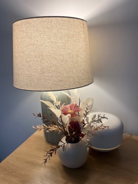 A table lamp with a beige fabric shade, a ceramic vase with pink and purple flowers and greenery, and a smart speaker on a wooden surface.