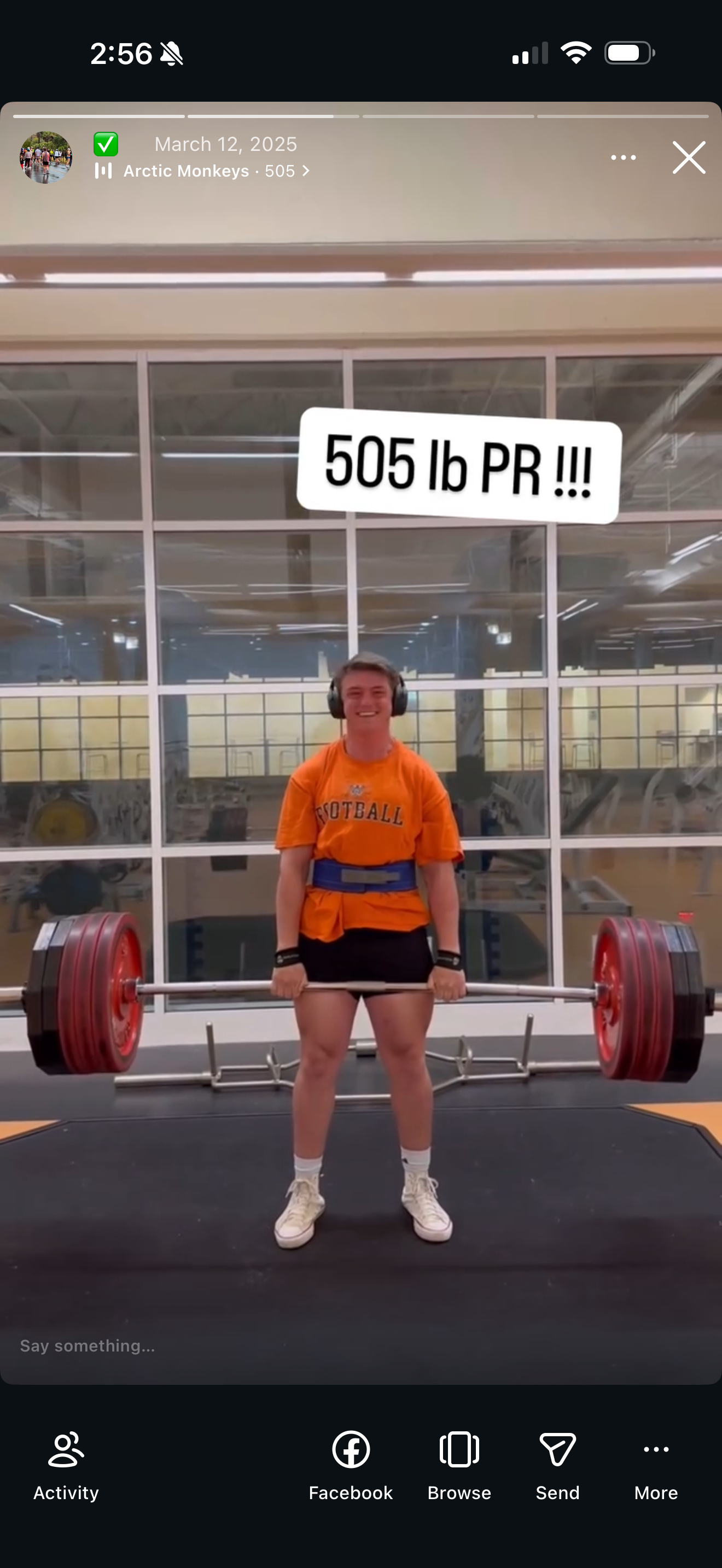 Young man in an orange football t-shirt and black shorts lifting a heavy barbell with red weight plates in a gym. He is smiling and wearing headphones, with a sign above him reading '505 lb PR!!!'