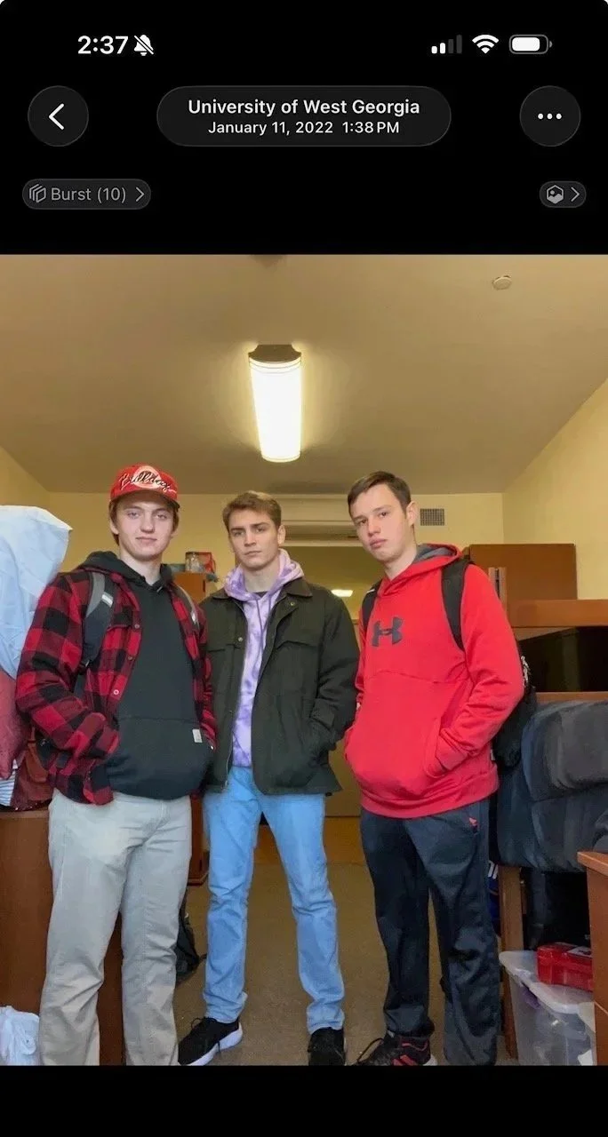 Three young men standing in a dorm room, dressed casually with hoodies, jackets, and backpacks, with a ceiling light overhead.
