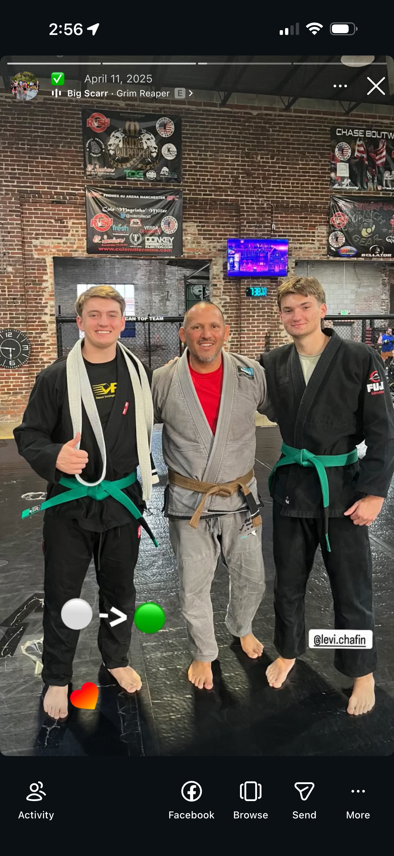 Two young male Brazilian Jiu-Jitsu practitioners with green belts and an instructor with brown belt, all in gi uniforms, standing together on a black mat inside a gym, smiling for a photo. The gym has brick walls and various banners hanging.