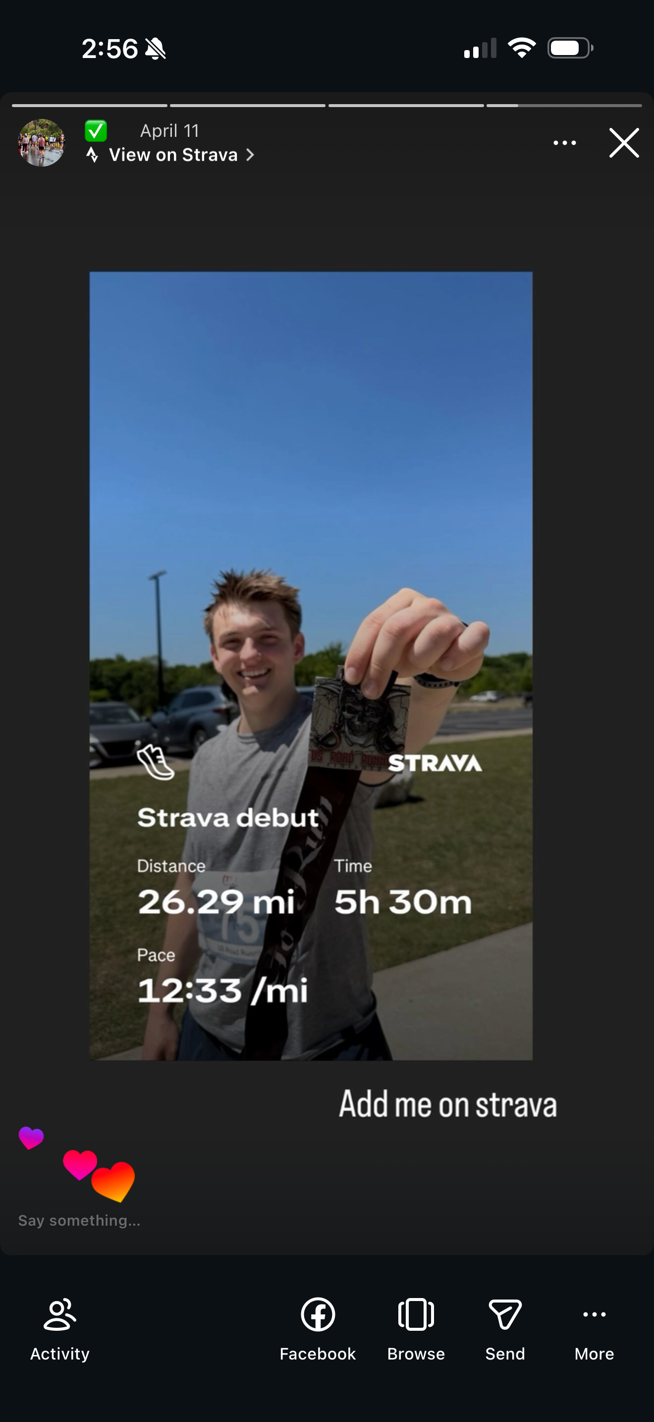 A young man outdoors on a sunny day, smiling and holding a medal and racing bib, with a parking lot and blue sky in the background.