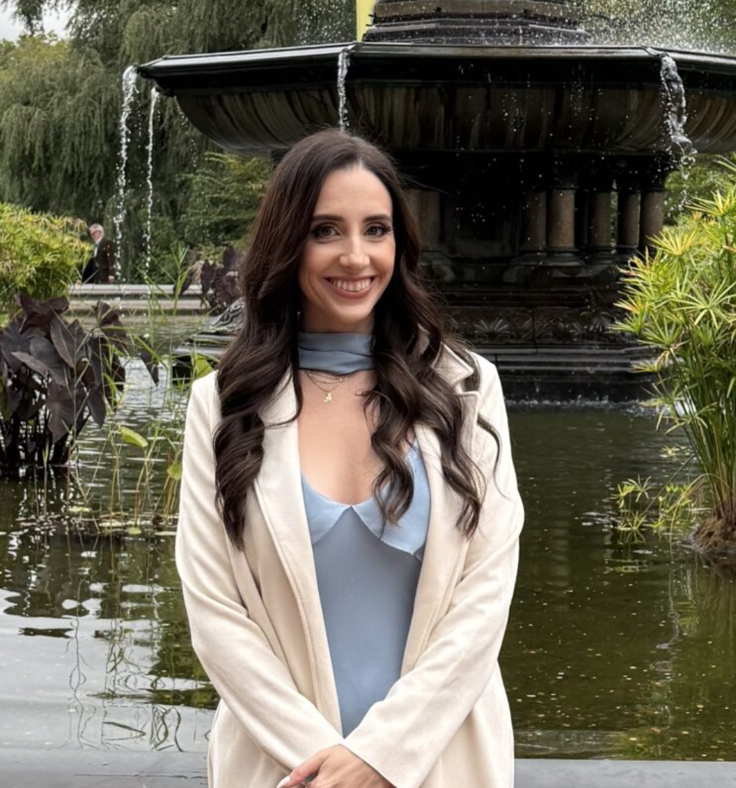 A young woman with long dark hair and a big smile standing in front of a fountain in a lush outdoor setting, wearing a cream-colored blazer over a light blue blouse.