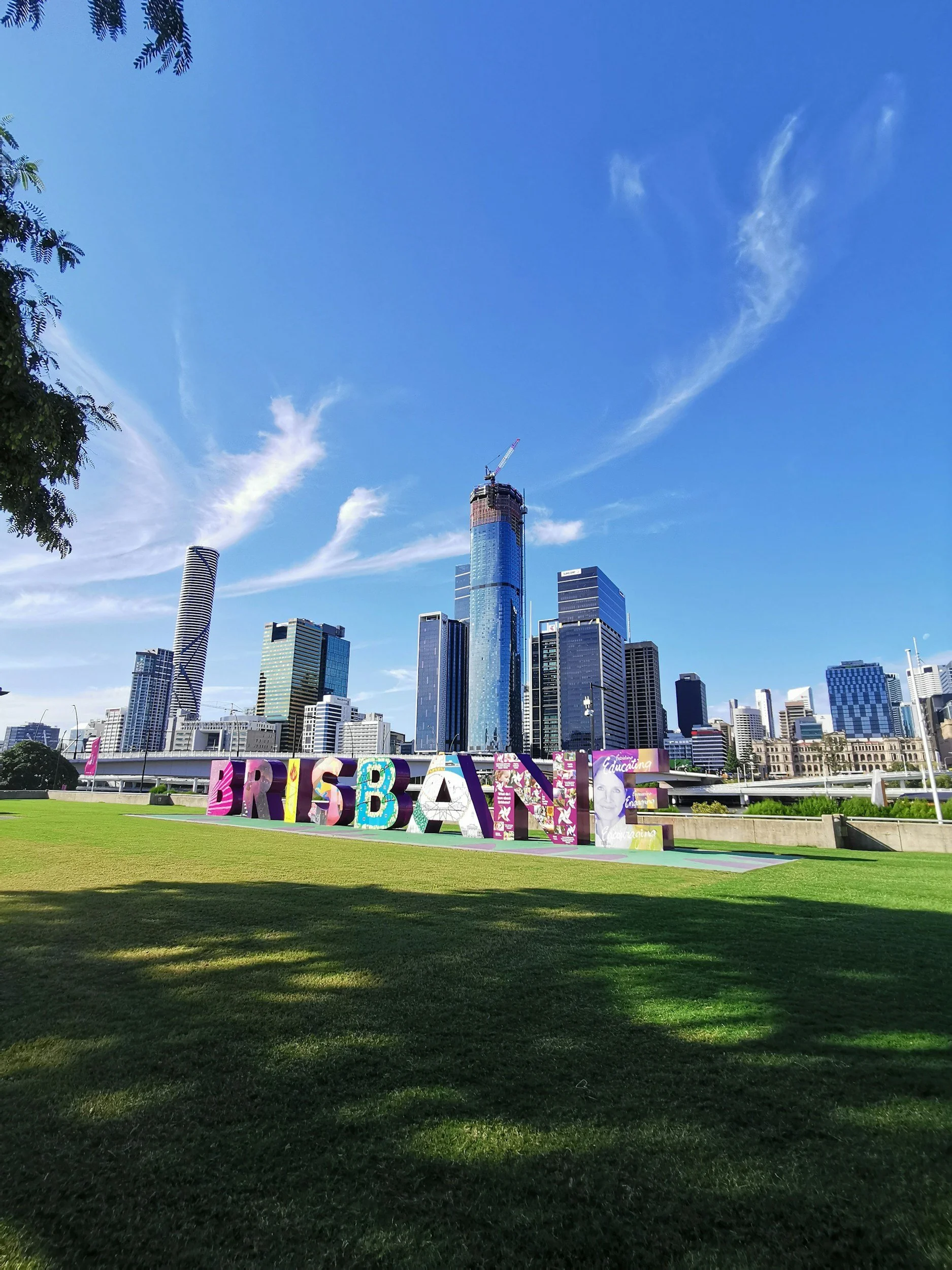 Colorful large letters spelling 'BRISBANE' in a park with a city skyline and blue sky in the background.