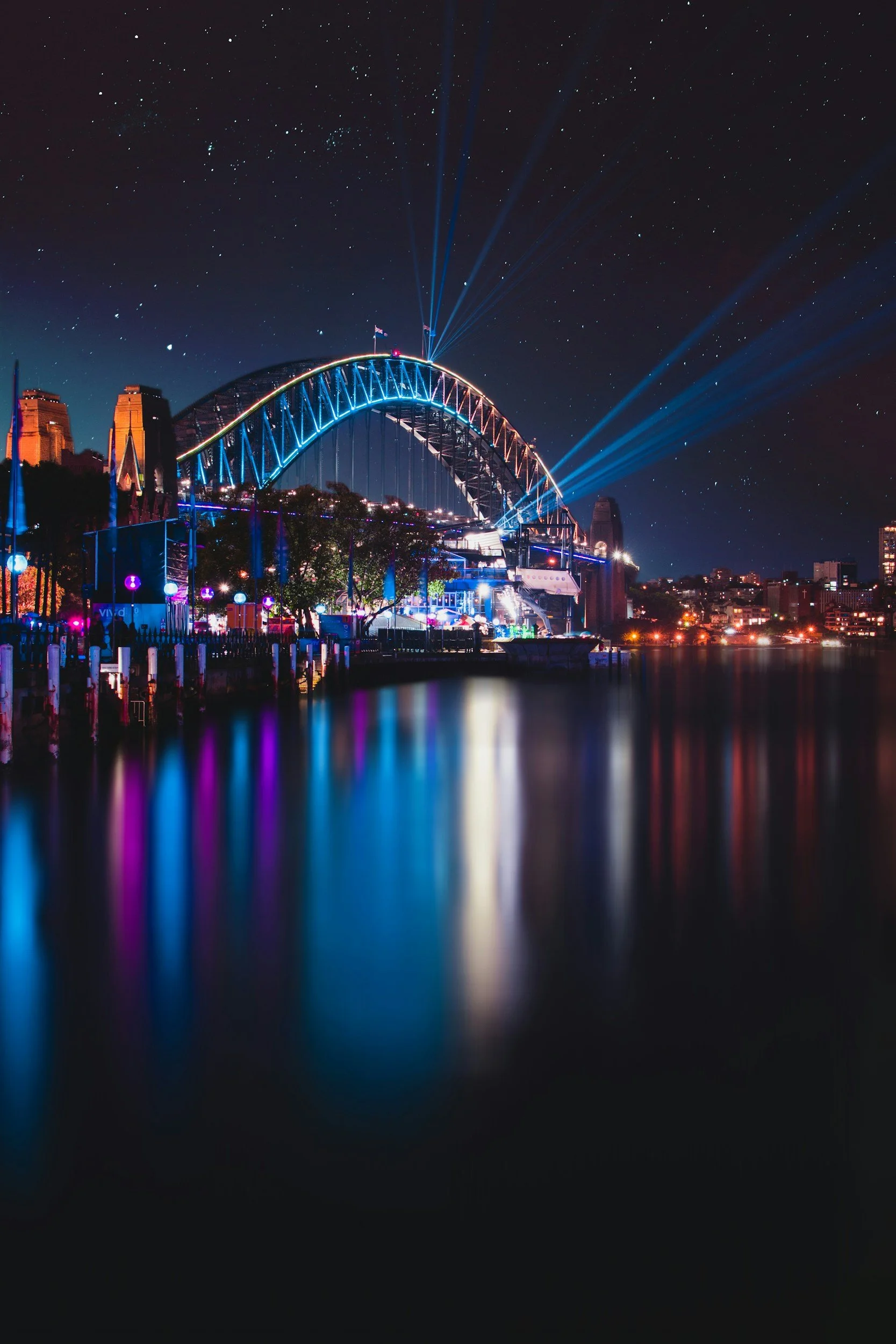 Night scene of a city skyline with a large illuminated bridge, fireworks, and colorful lights reflecting on the water below.