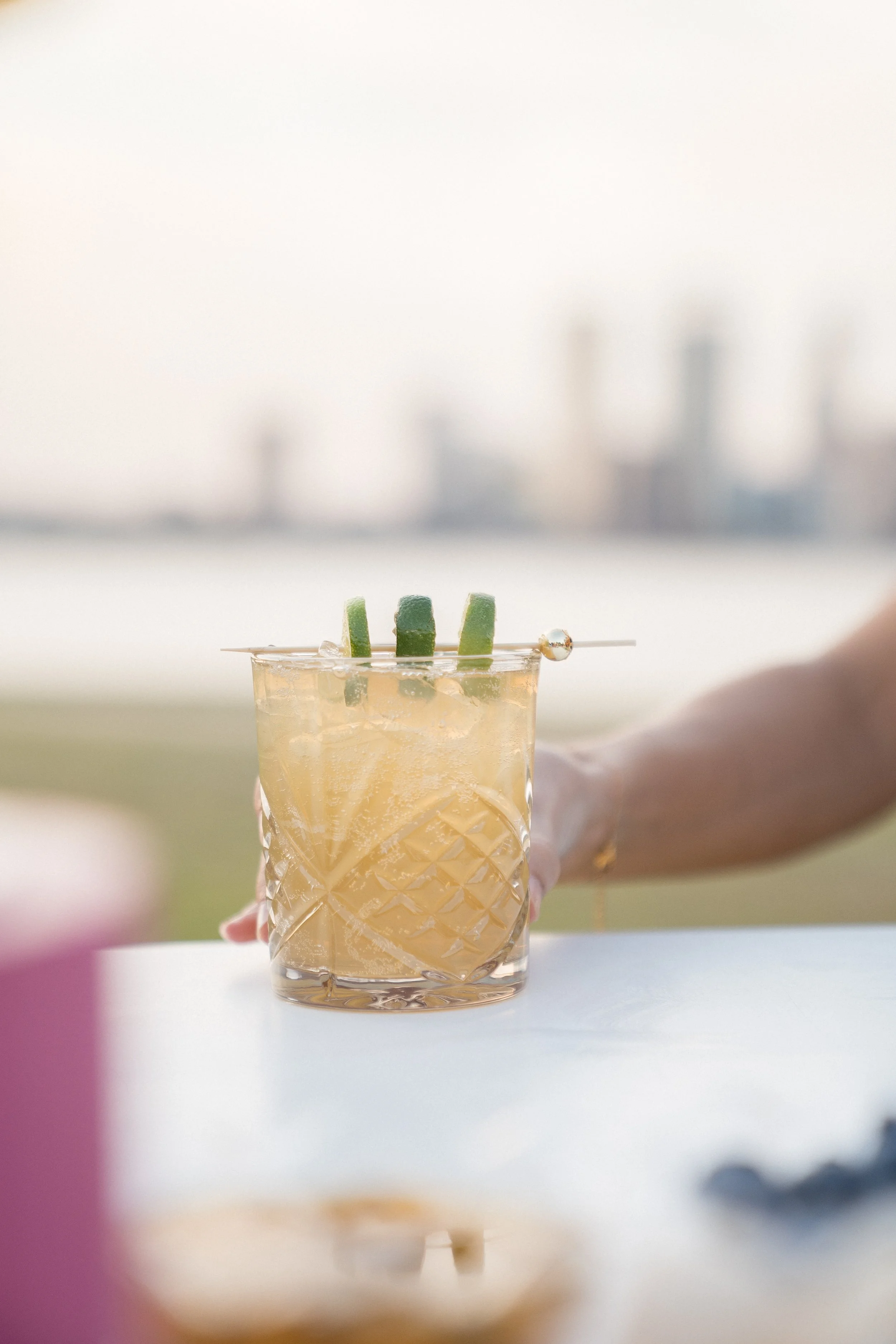 A hand holding a glass of iced tea with slices of cucumber garnishing the drink, with a blurred background of a city skyline.