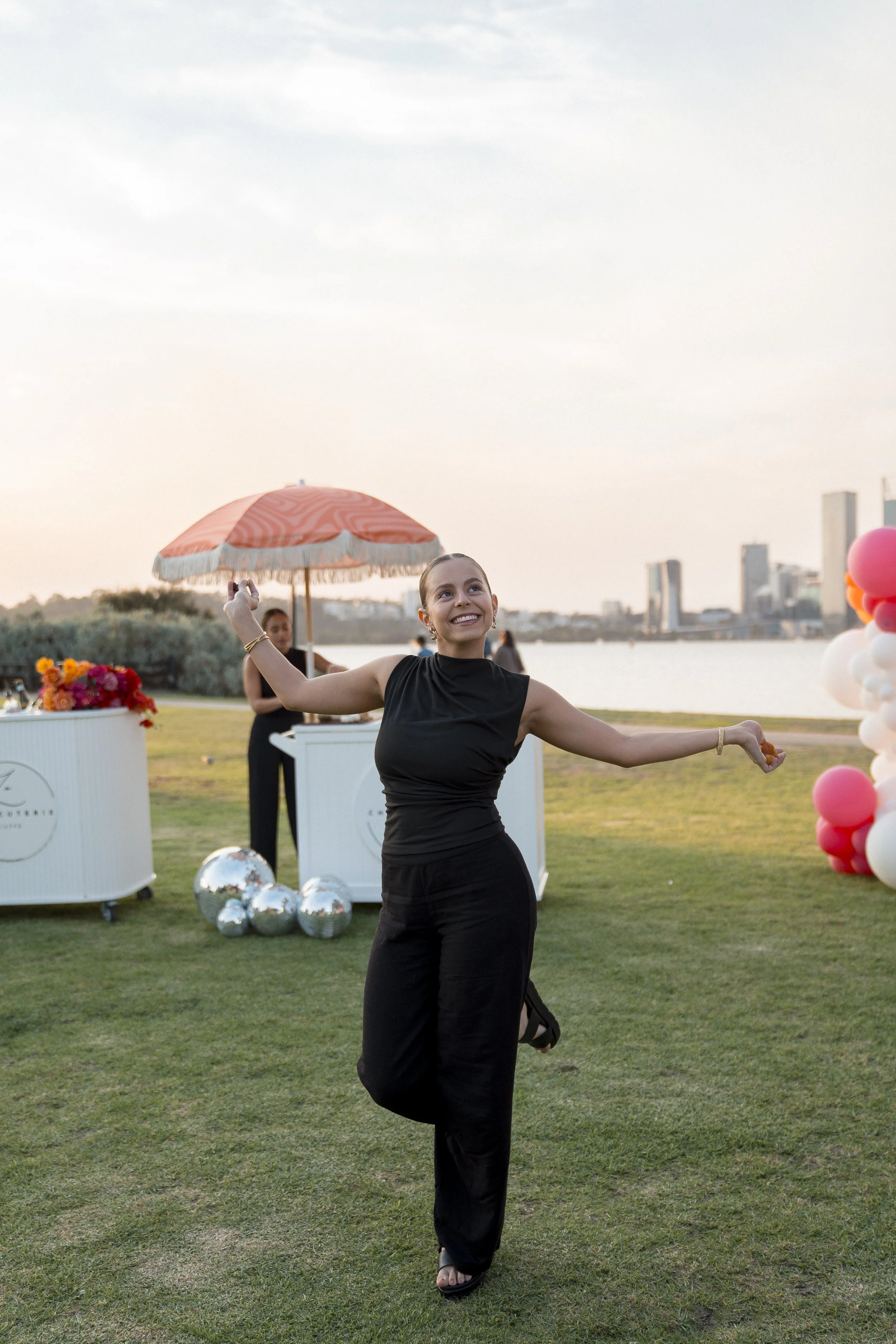 A woman in a black dress dancing outdoors near a river with a city skyline in the background. There are balloons, flowers, and reflective spheres around her, and a woman under an orange parasol in the background.