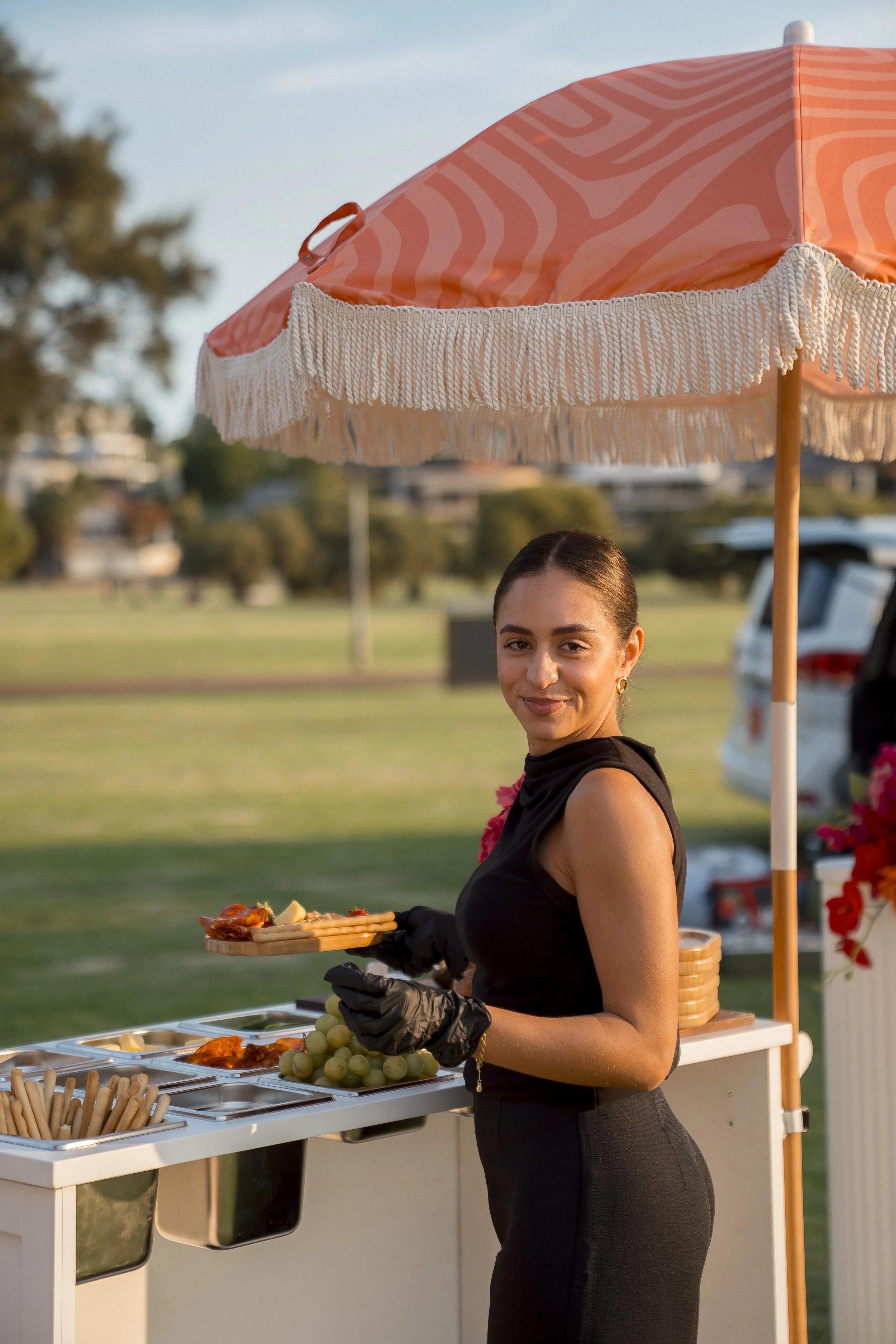 A woman in black dress serving food at an outdoor buffet table with a large pink umbrella providing shade