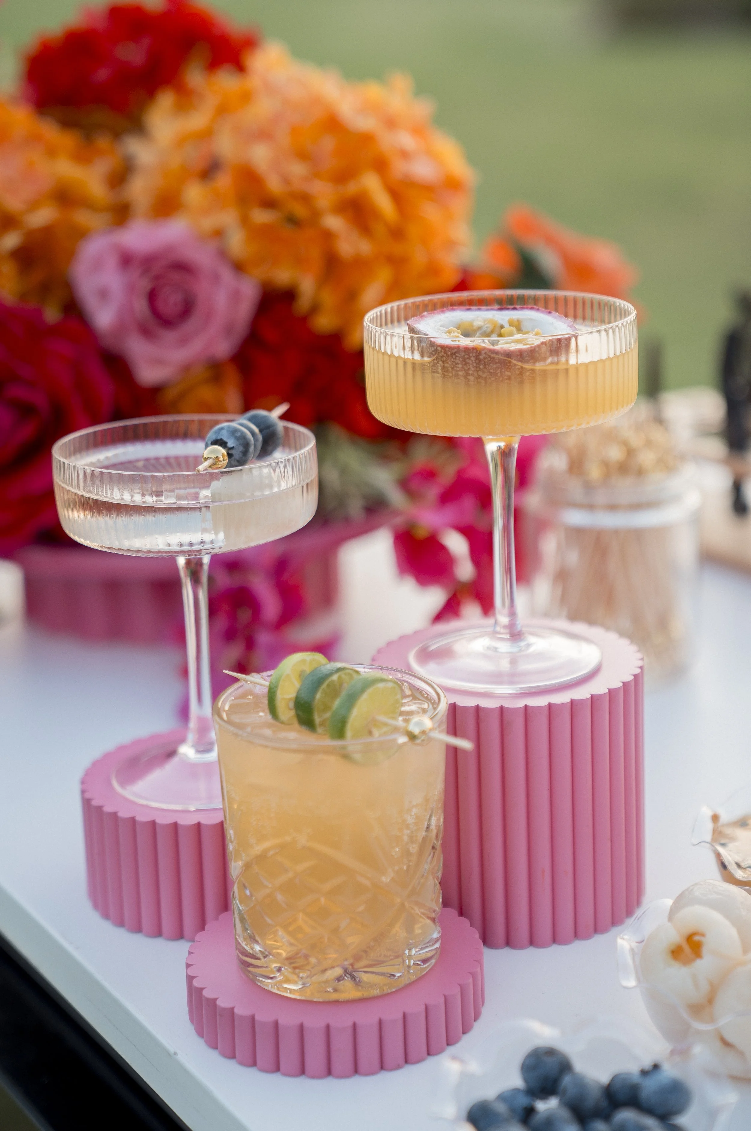 Presentation of three colorful cocktails on pink platforms, with a blurred bouquet of flowers in the background and blueberries in the foreground.