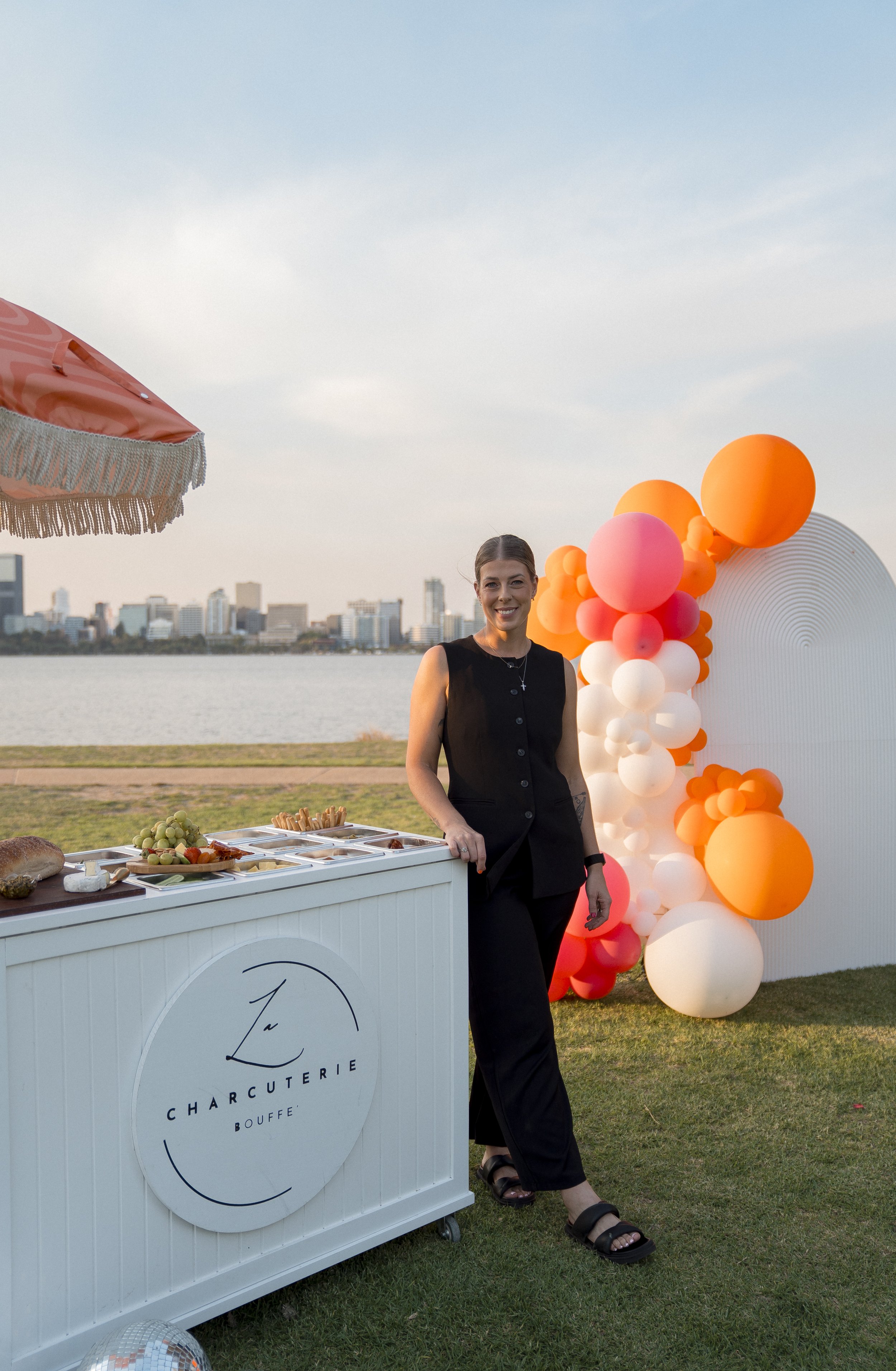 A woman standing next to a white charcuterie stand with a city skyline in the background, decorated with orange, pink, and white balloons, during sunset.