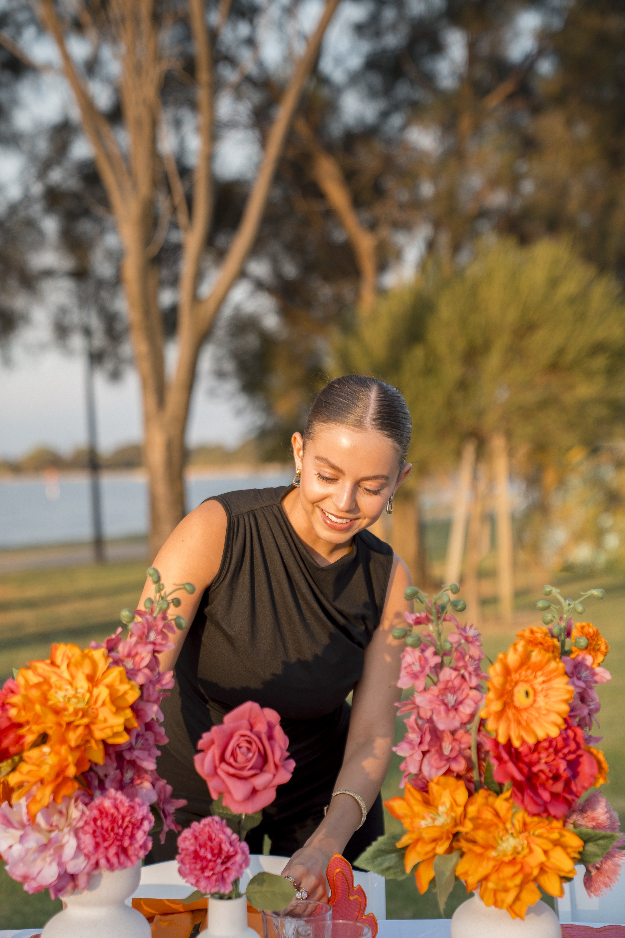 A woman in a black dress arranging colorful flowers on a table outdoors during sunset, with trees and a body of water in the background.