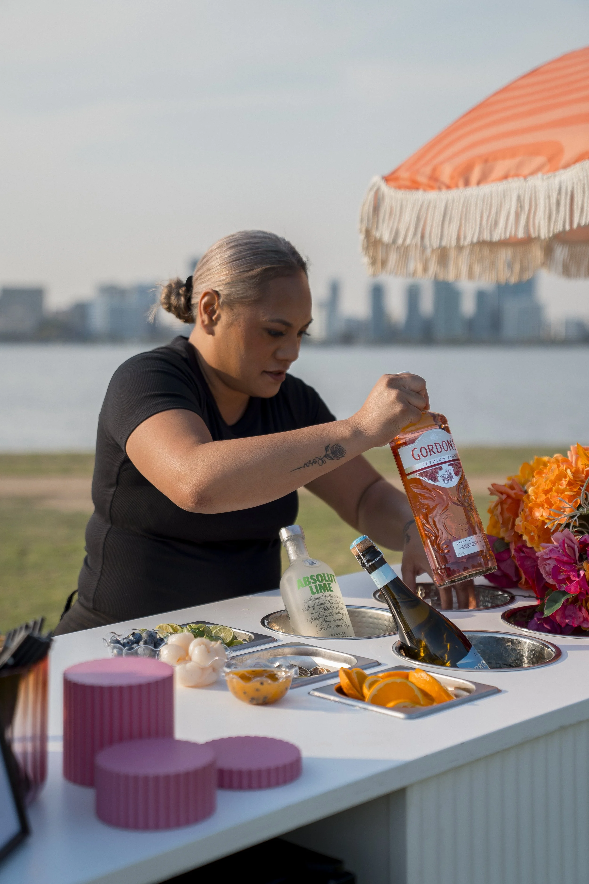 A woman with gray hair tied in a bun opening a bottle of Gordon's gin at an outdoor event with a city skyline in the background. The table has various food items, flowers, and liquor bottles, with a large orange and cream umbrella overhead.