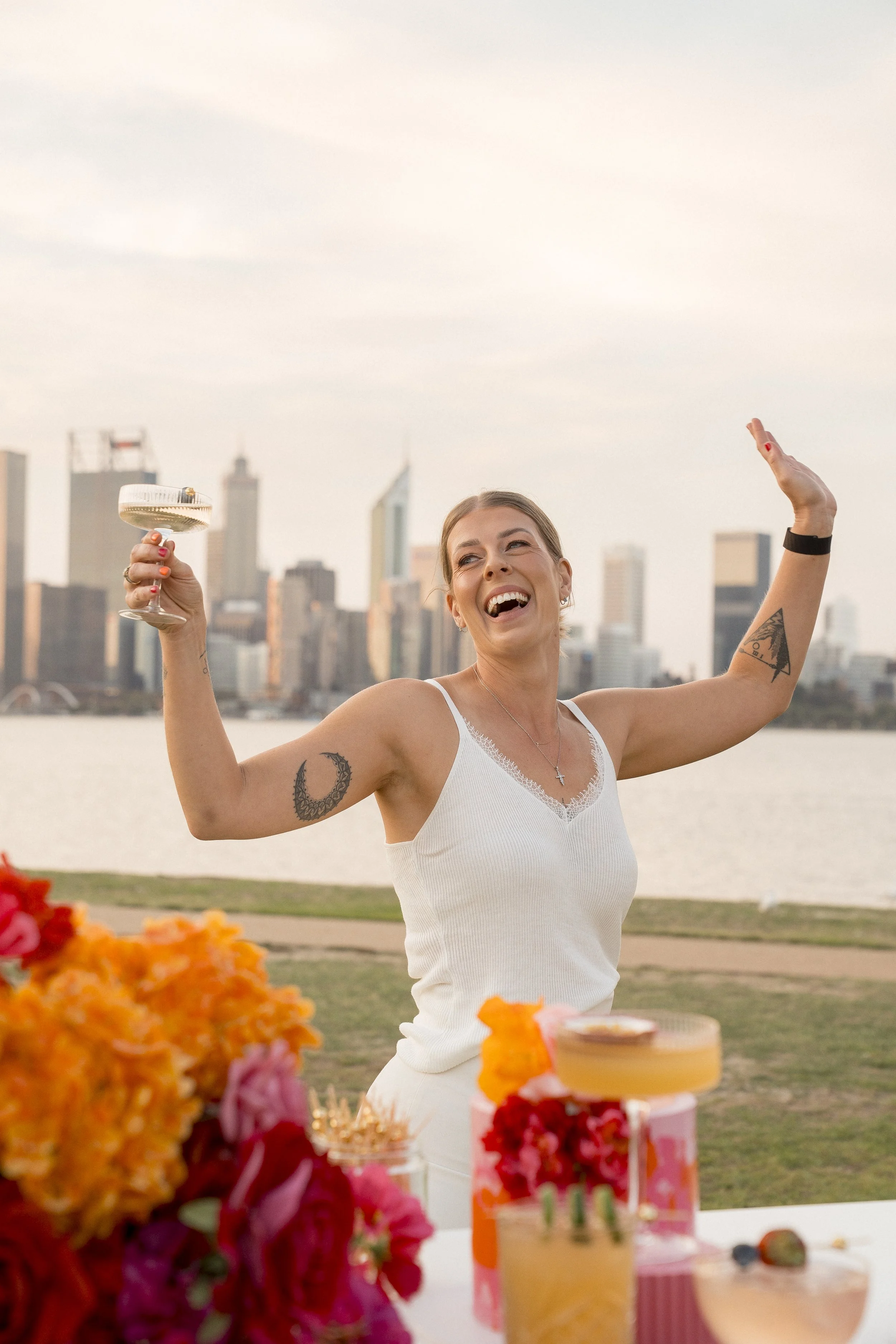 A woman at an outdoor celebration holding a glass of champagne, with a city skyline in the background and colorful flowers and desserts in the foreground.