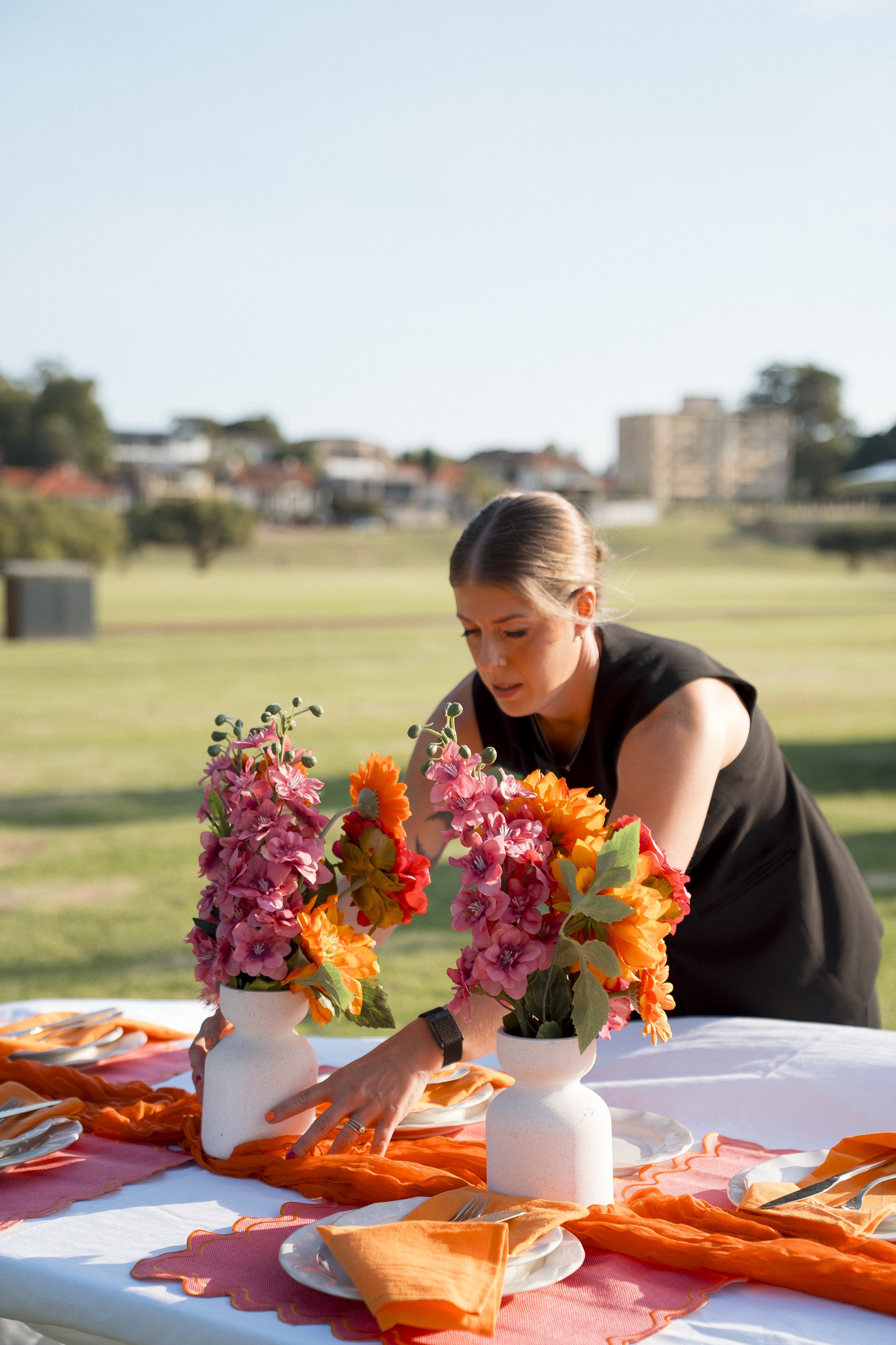 A woman arranging flower arrangements on a table outdoors with a cityscape in the background.