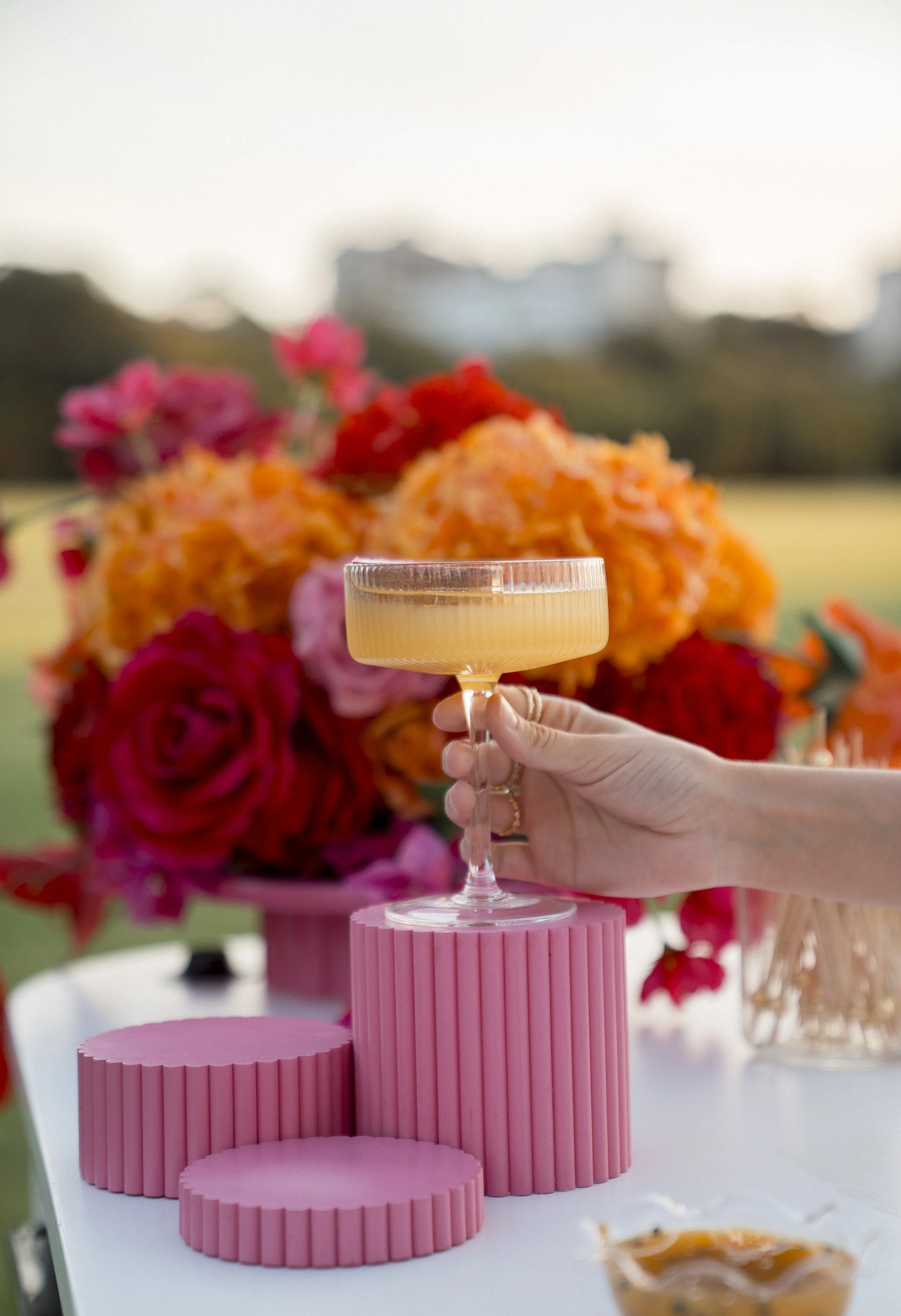Person holding a champagne flute with a pink and orange floral arrangement in the background and pink tiered stands on a table.