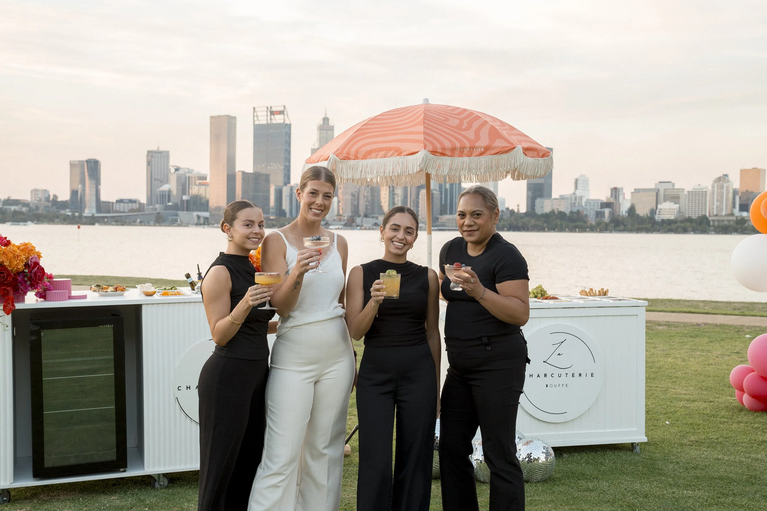 Four women standing together outdoors near a body of water, holding drinks, with a city skyline in the background and a pink and orange beach umbrella overhead.