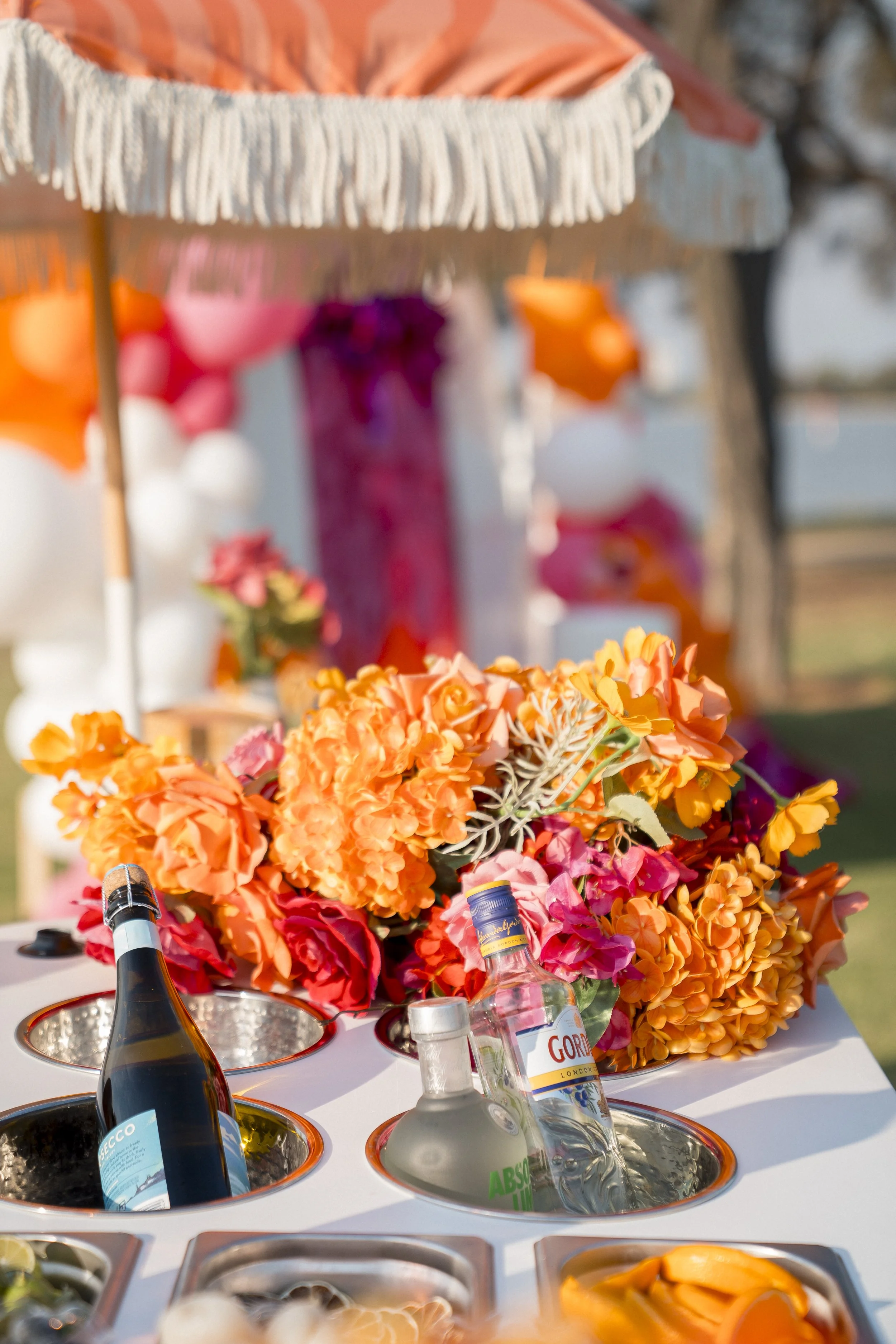 A colorful floral centerpiece surrounded by bottles and drink supplies on a table at an outdoor event.