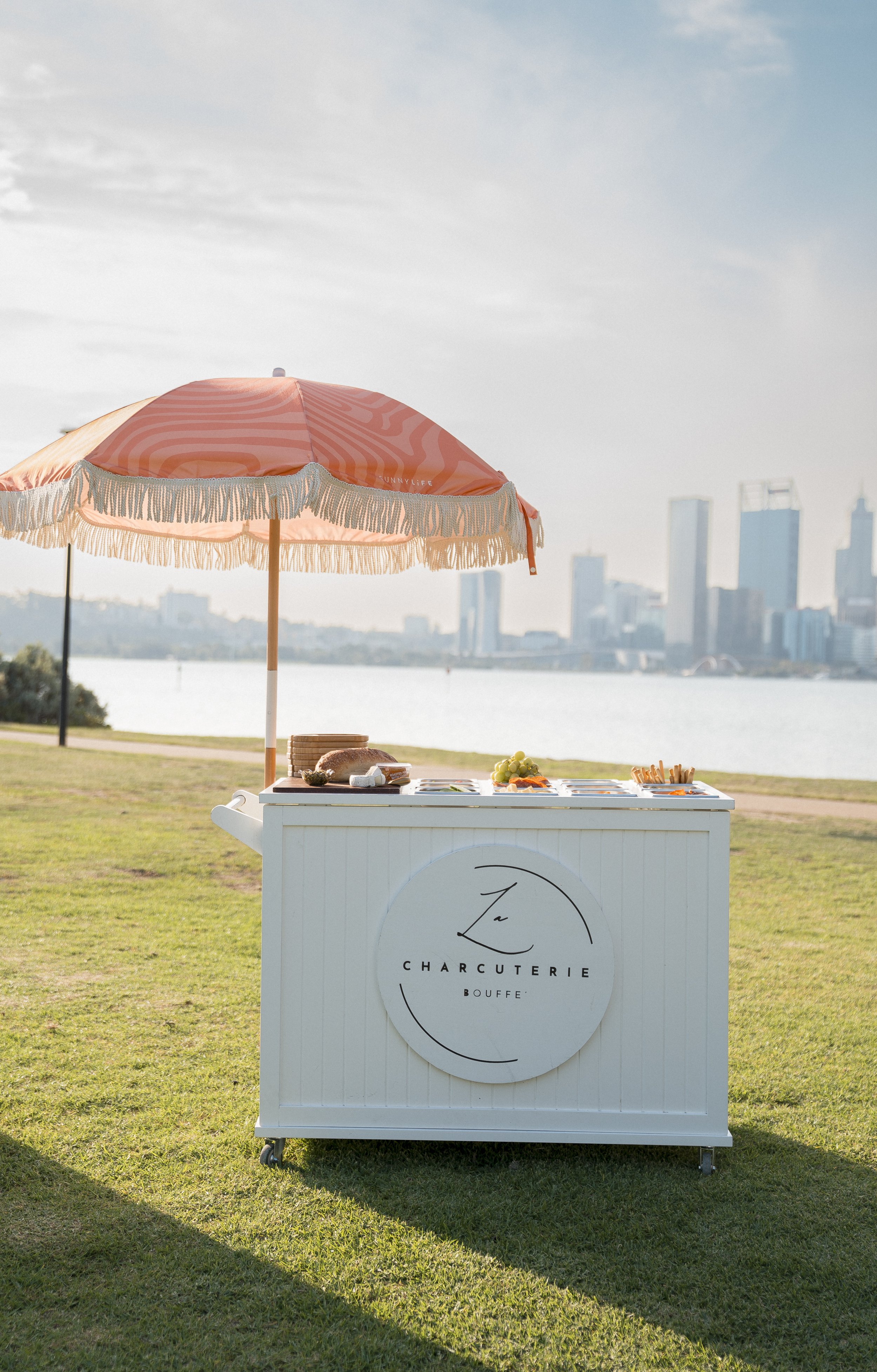 A white charcuterie food stand with a circular logo reading 'La Charcuterie Bouffe' on the front, set on grass near a body of water with a city skyline in the background. There is a large pink and beige umbrella providing shade over the stand, which 