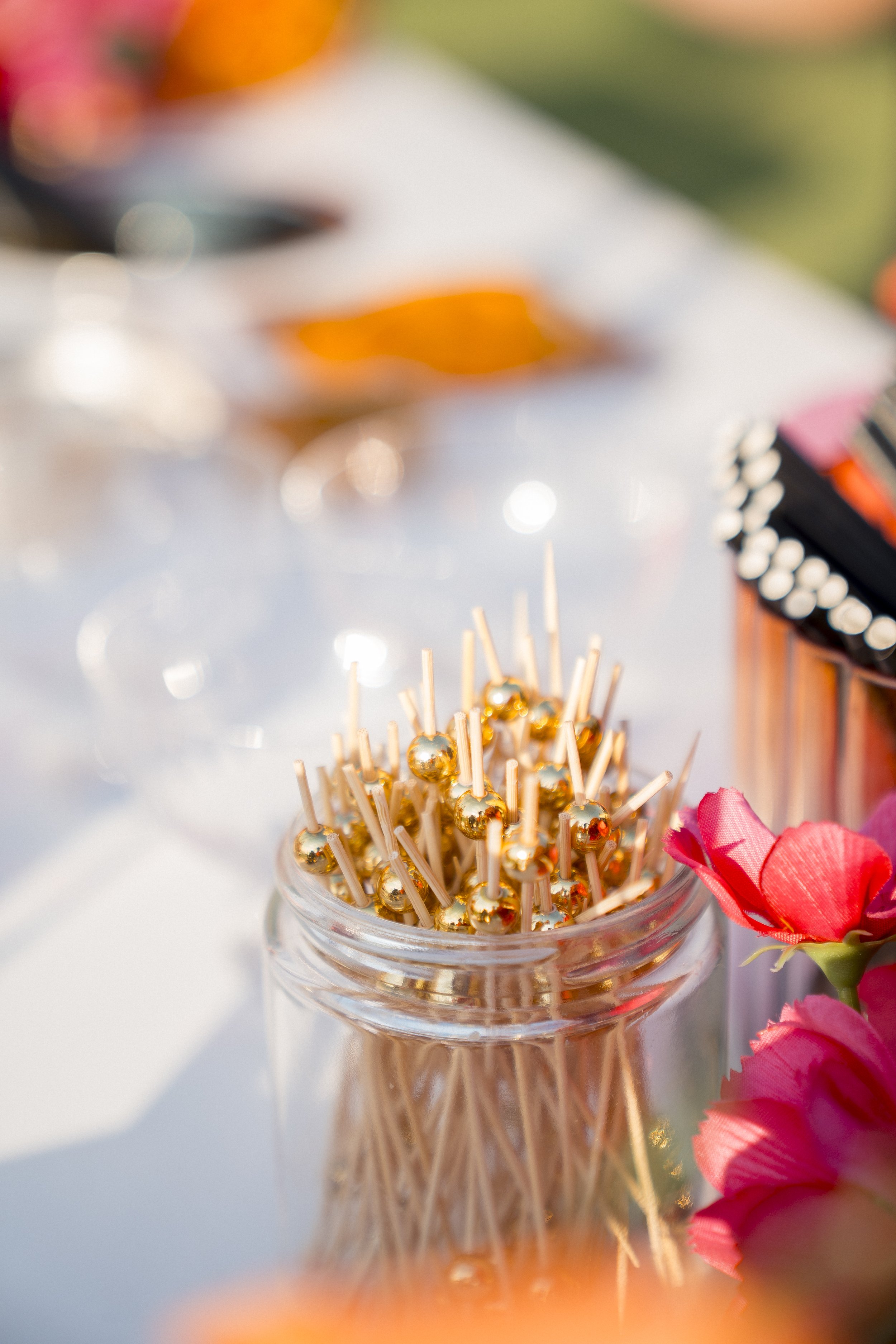 Close-up of a glass jar filled with gold-colored toothpick holders with reflective balls on top, next to pink flowers, on a white table with colorful and blurred background.