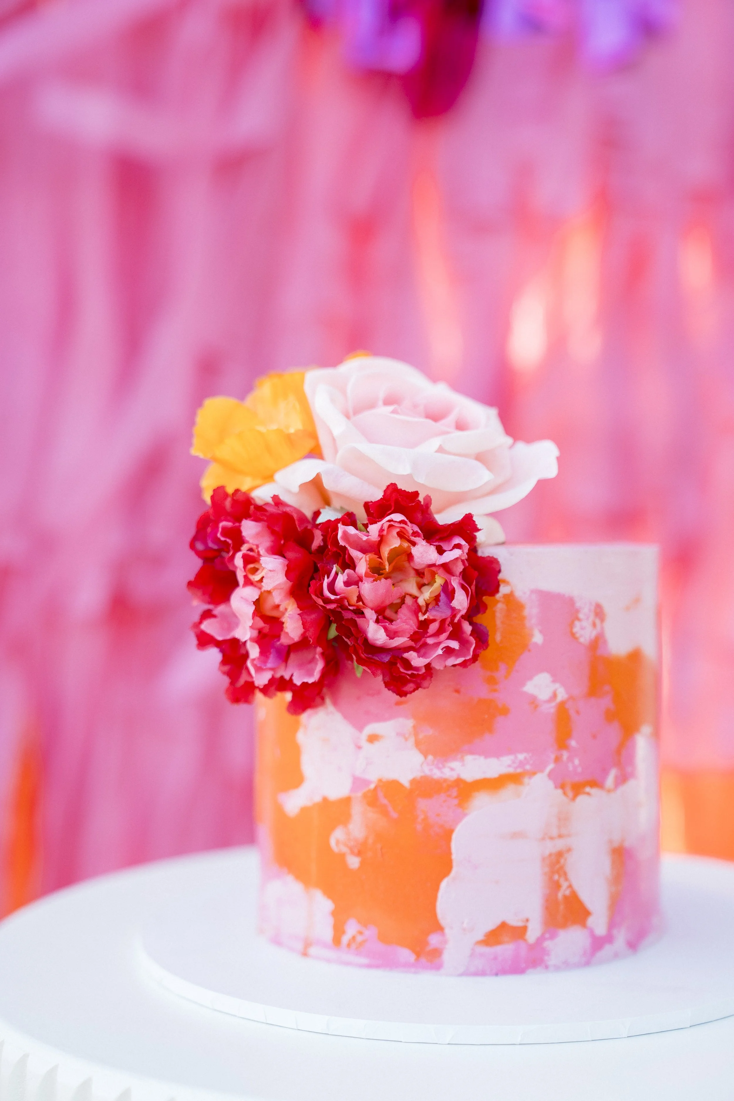 A pink and white marbled cake with floral decorations, including a pink rose, yellow petals, and red flowers, on top.
