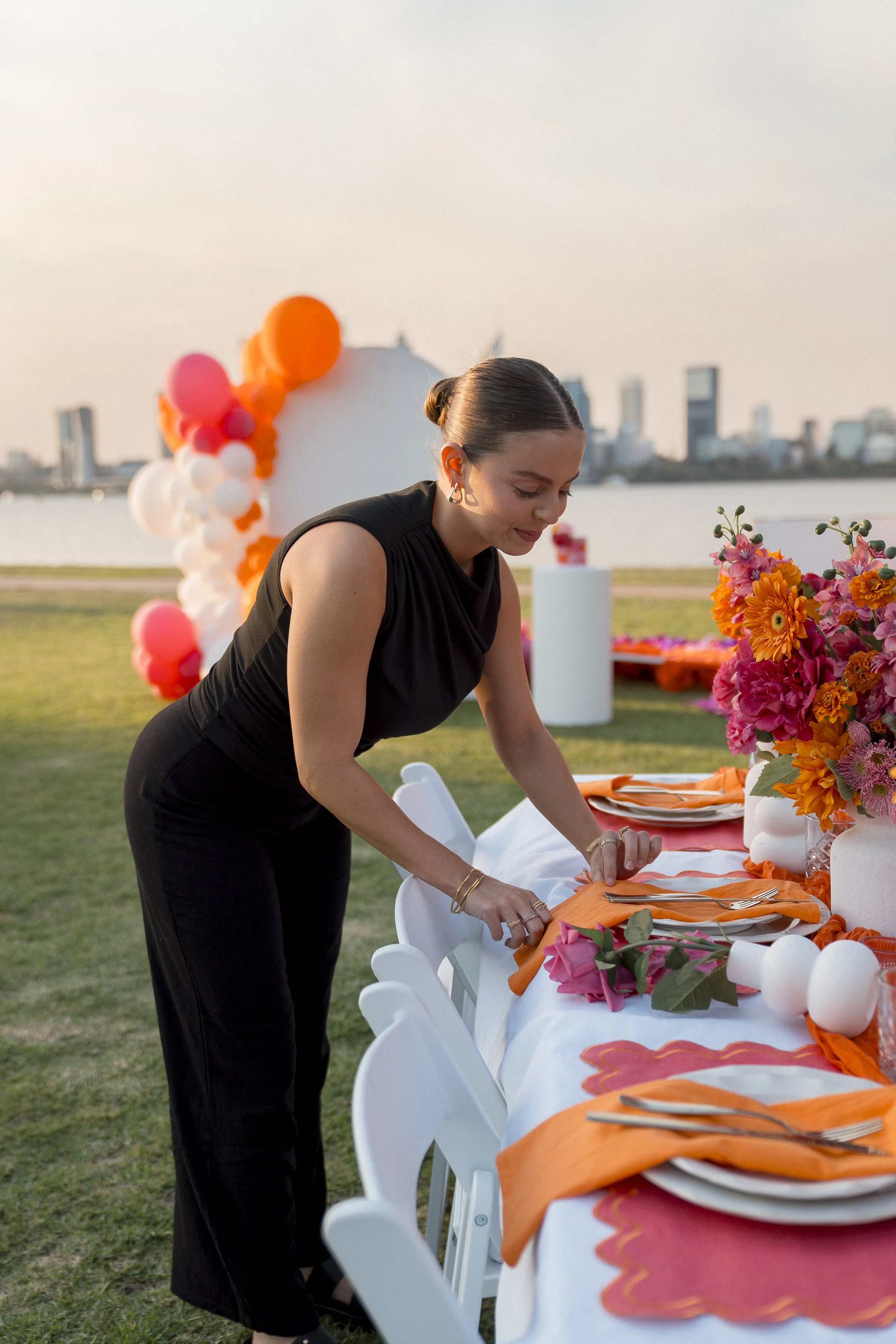 A woman in a black dress is setting a long outdoor dining table decorated with orange, pink, and white floral arrangements, with balloons in similar colors in the background, set near a body of water during sunset with a city skyline in the distance.
