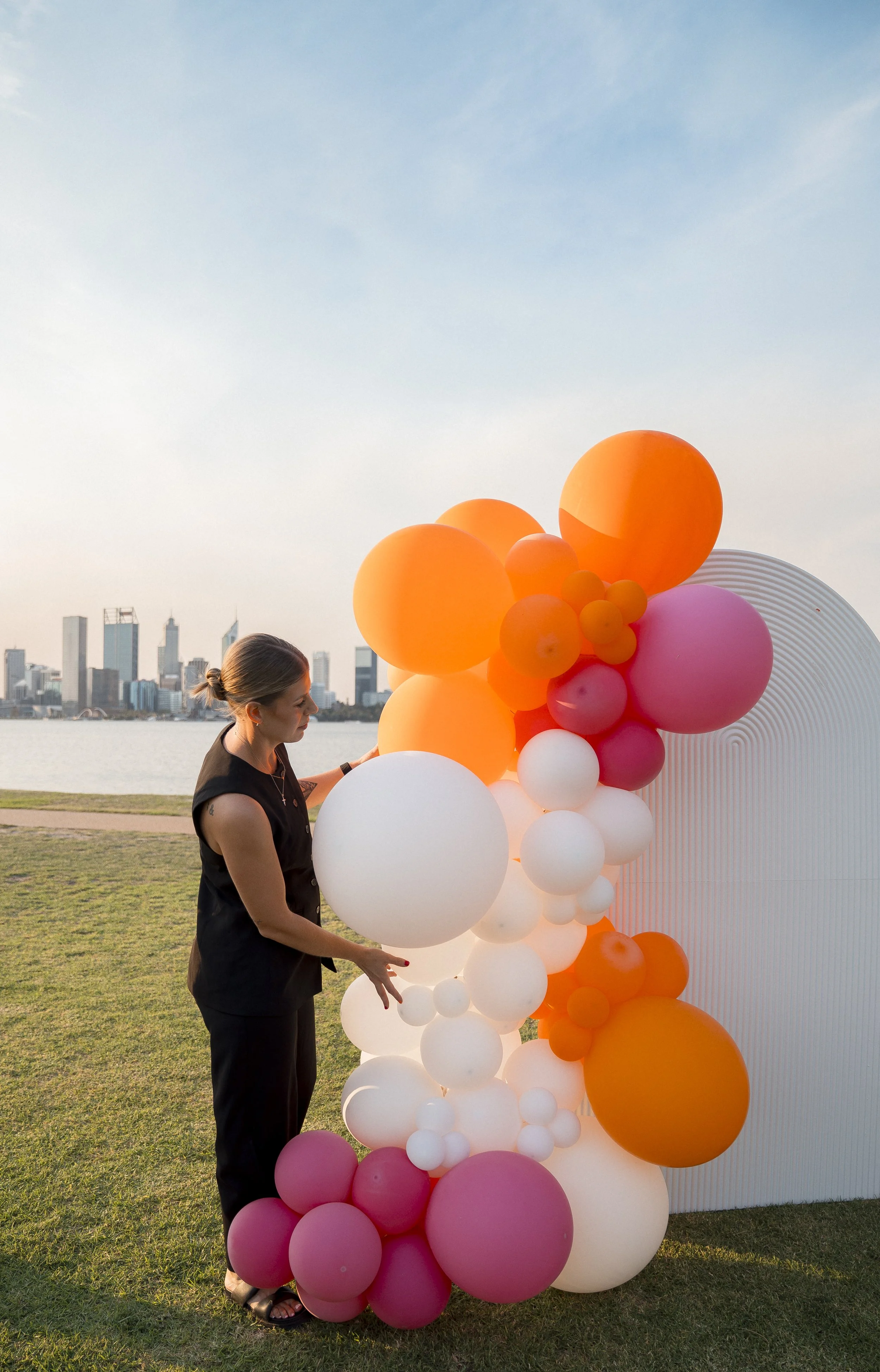 A woman standing outdoors near a colorful balloon sculpture with a city skyline in the background during sunset.