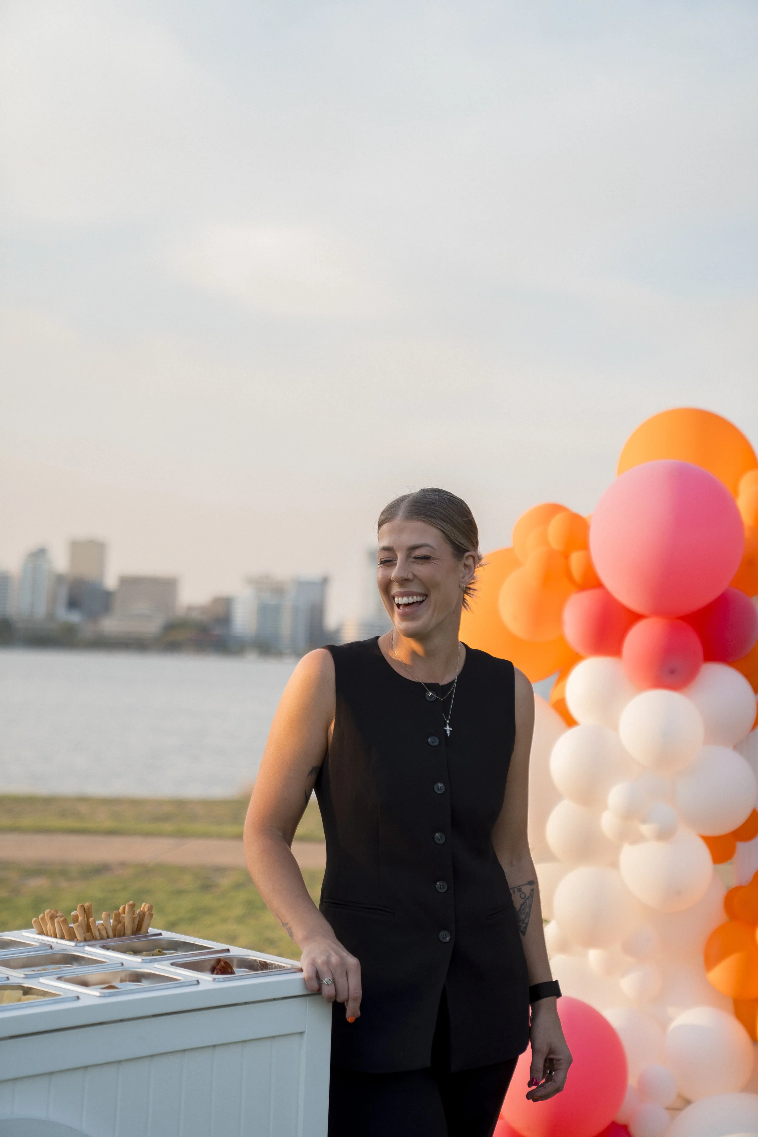 A smiling woman in a black sleeveless dress standing outdoors near a balloon decoration in pink, orange, and white colors, with a cityscape and water in the background during the evening.
