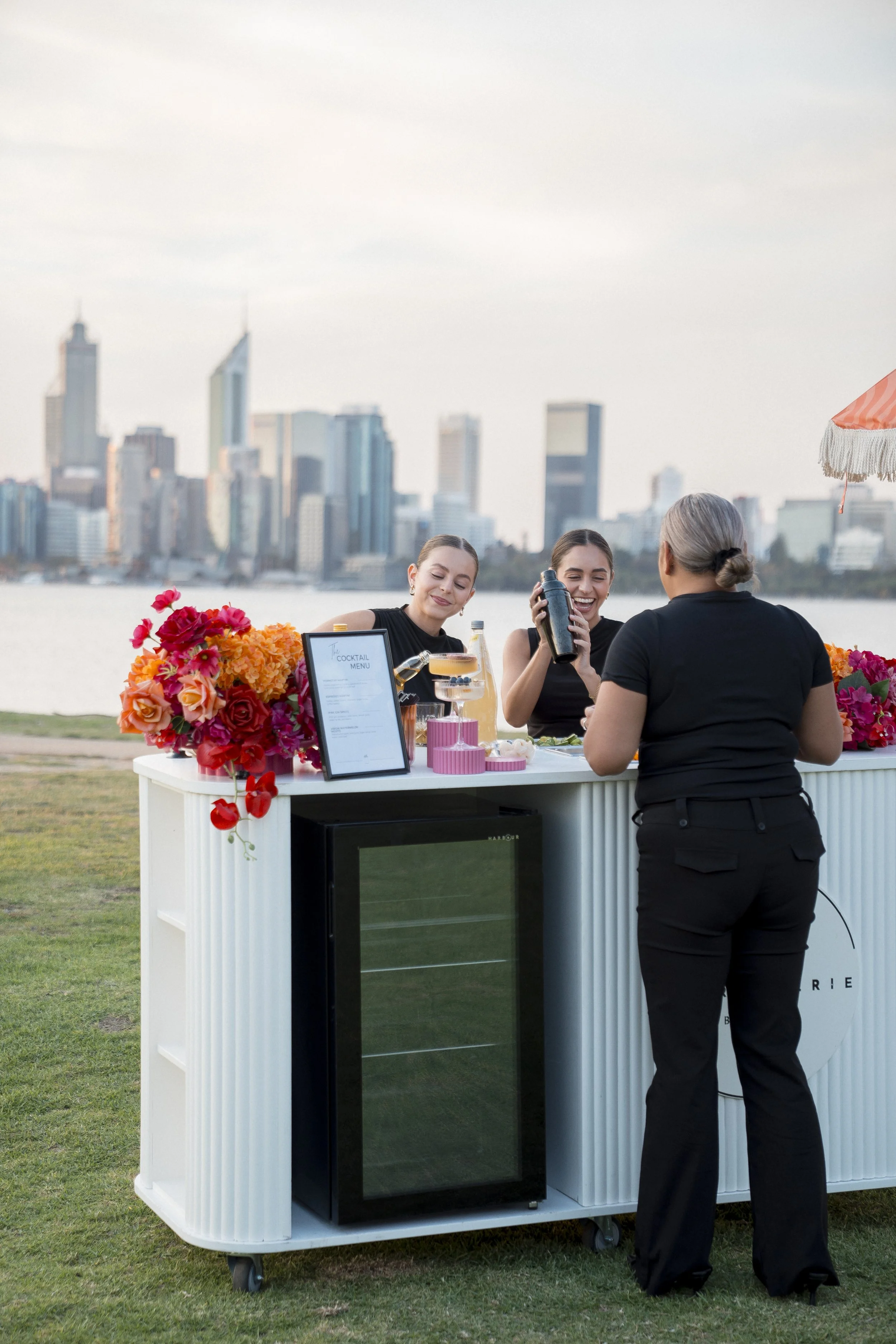 A daytime outdoor scene with two women sitting at a table decorated with colorful flowers, and a third woman standing in front of a portable bar in front of the water with a city skyline in the background. The women are smiling and holding drinks.