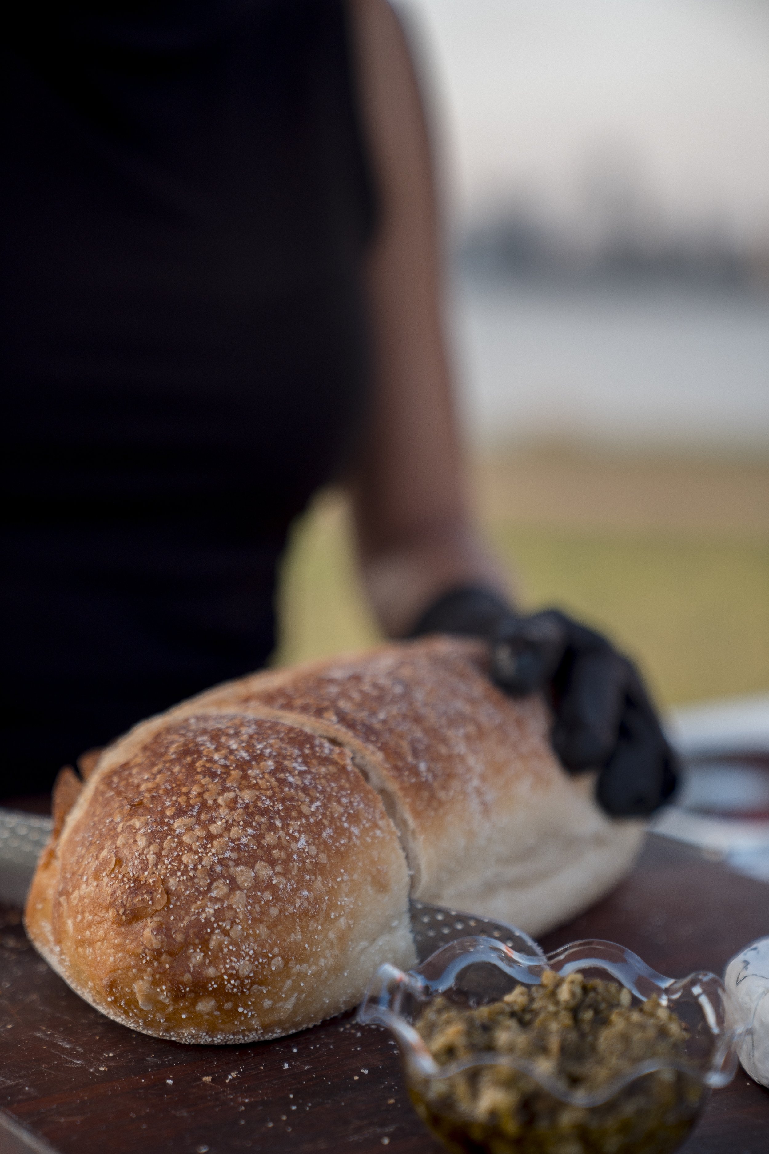 Close-up of a toasted hot dog bun with salt crystals on top, placed on a dark wooden surface, with a small glass bowl of relish and some spilled salt crystals nearby, and a blurred background including a person in black attire.