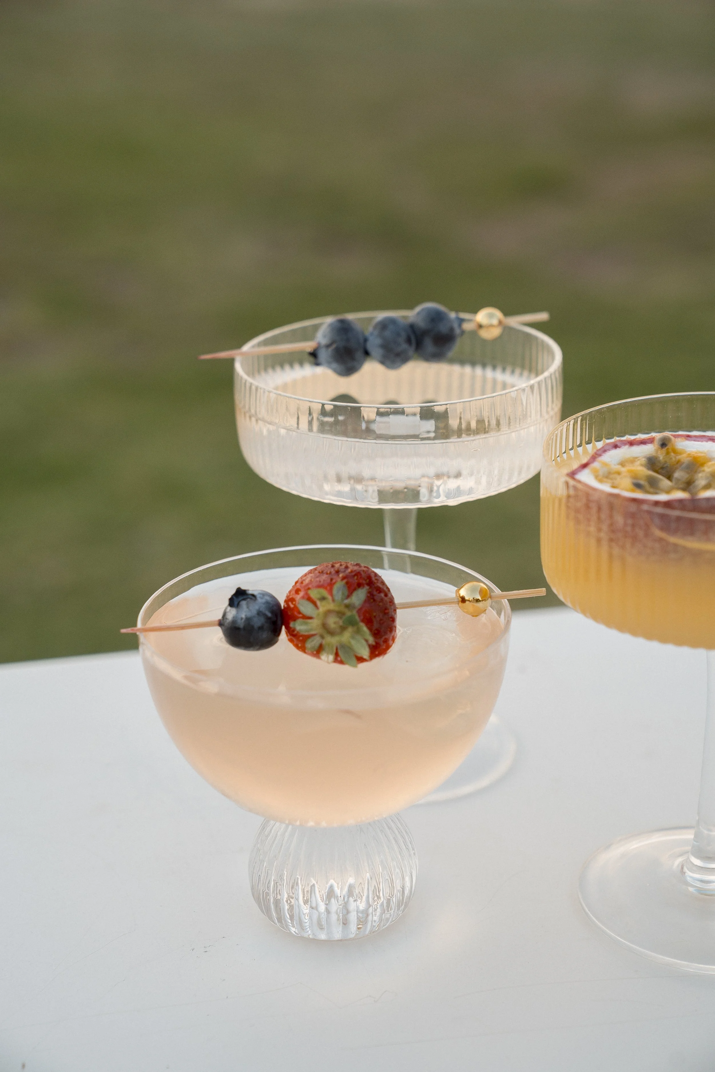 A pink cocktail with blueberries and a strawberry garnished with a small gold ball and toothpick, served in a vintage glass. Two other similar drinks are partially visible, one with blueberries and a gold bead, and another with a yellowish hue and fr