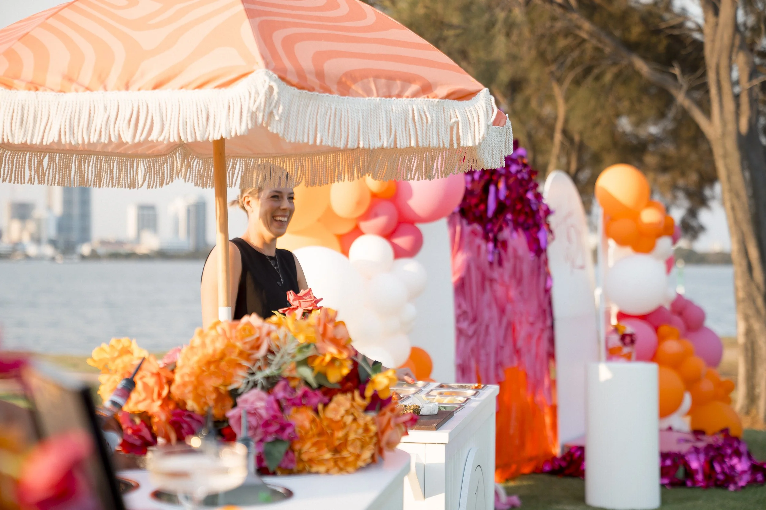 A smiling woman standing behind a colorful flower-covered booth at an outdoor event, with balloons and decorations in the background near a body of water and trees.