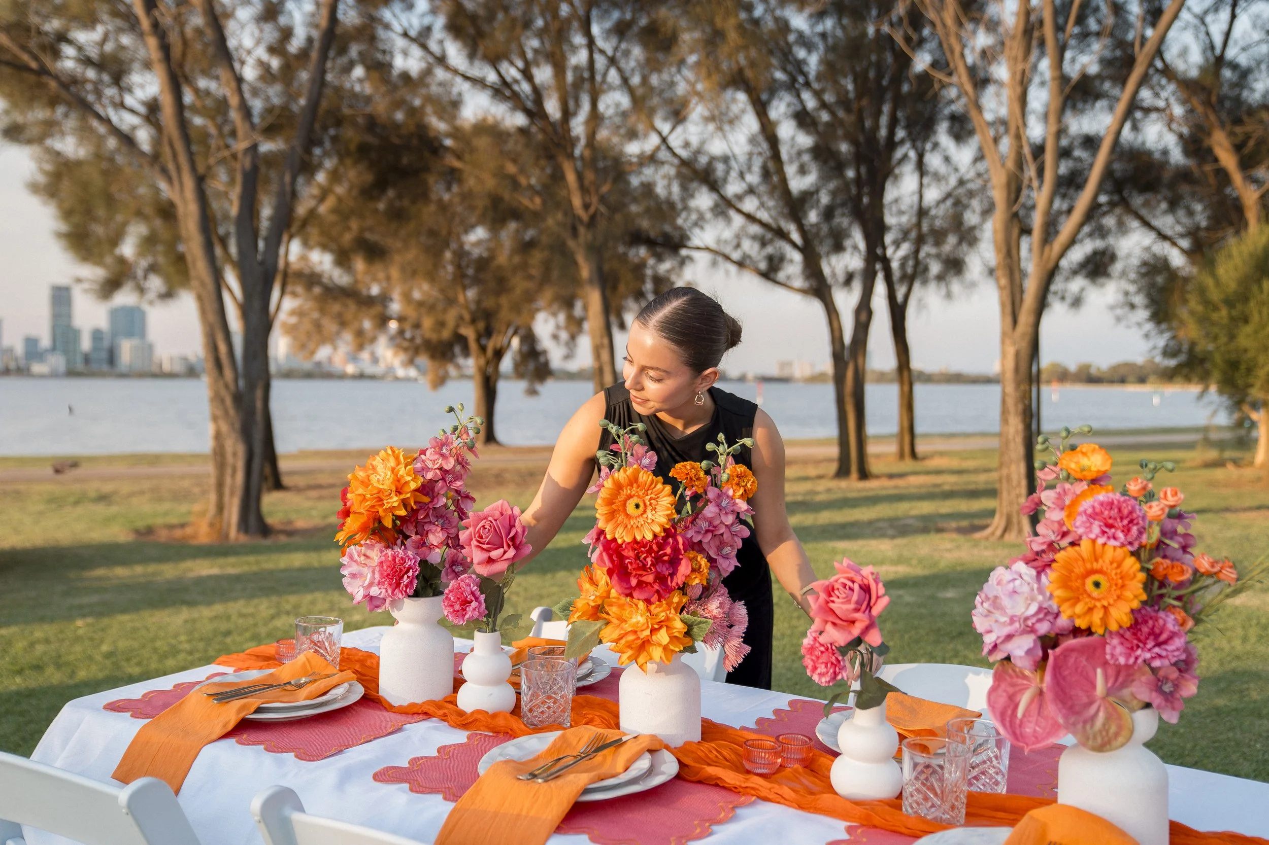 A woman setting a table with floral arrangements outdoors near a water body, with trees and a city skyline in the background during sunset.