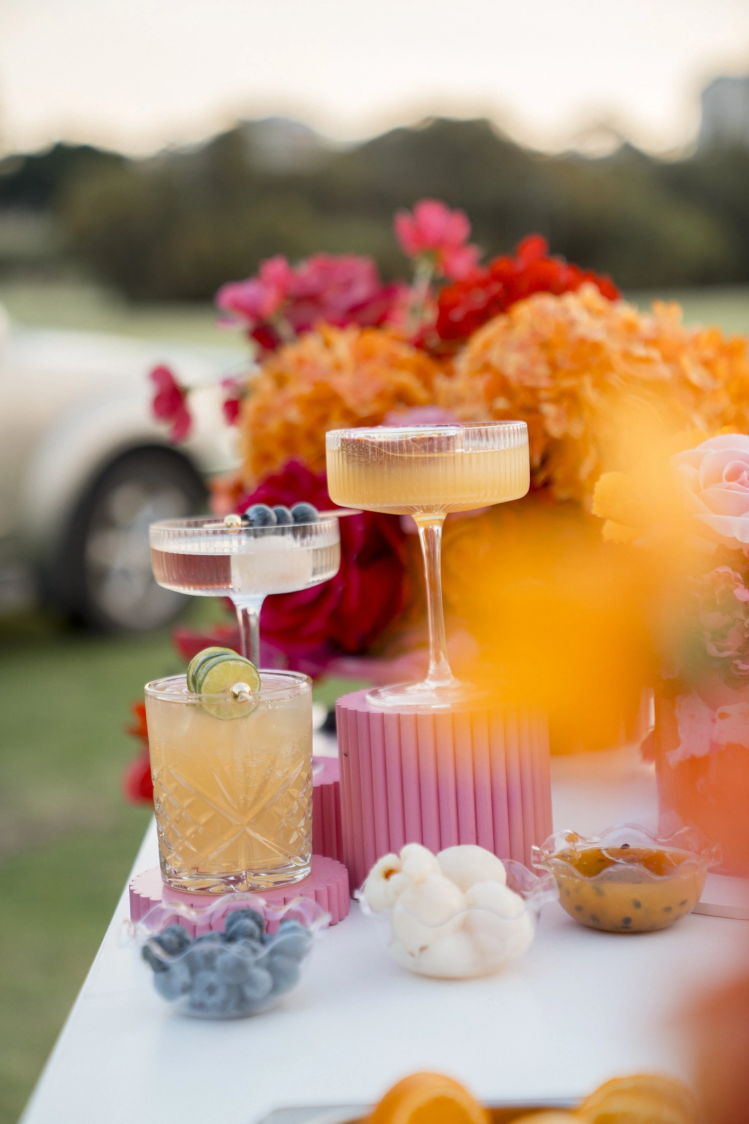 A table with various colorful desserts and drinks, including a margarita with blueberries and lemon, surrounded by flowers and decorative elements.