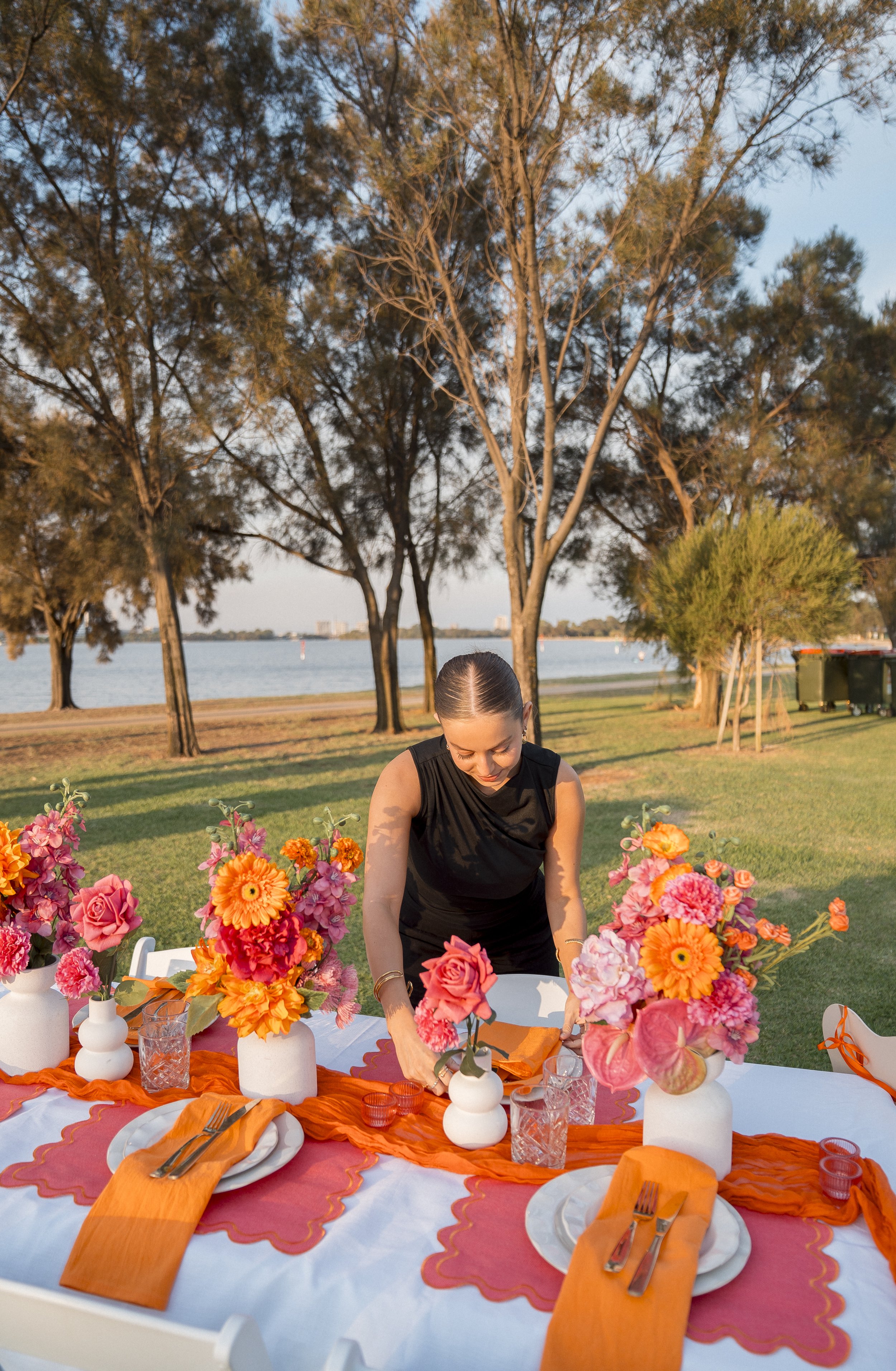 A woman is setting a table outdoors for a celebration, with a background of trees, water, and a clear sky at sunset. The table is decorated with orange and pink flowers in white vases, and orange table runners with place settings and glassware.