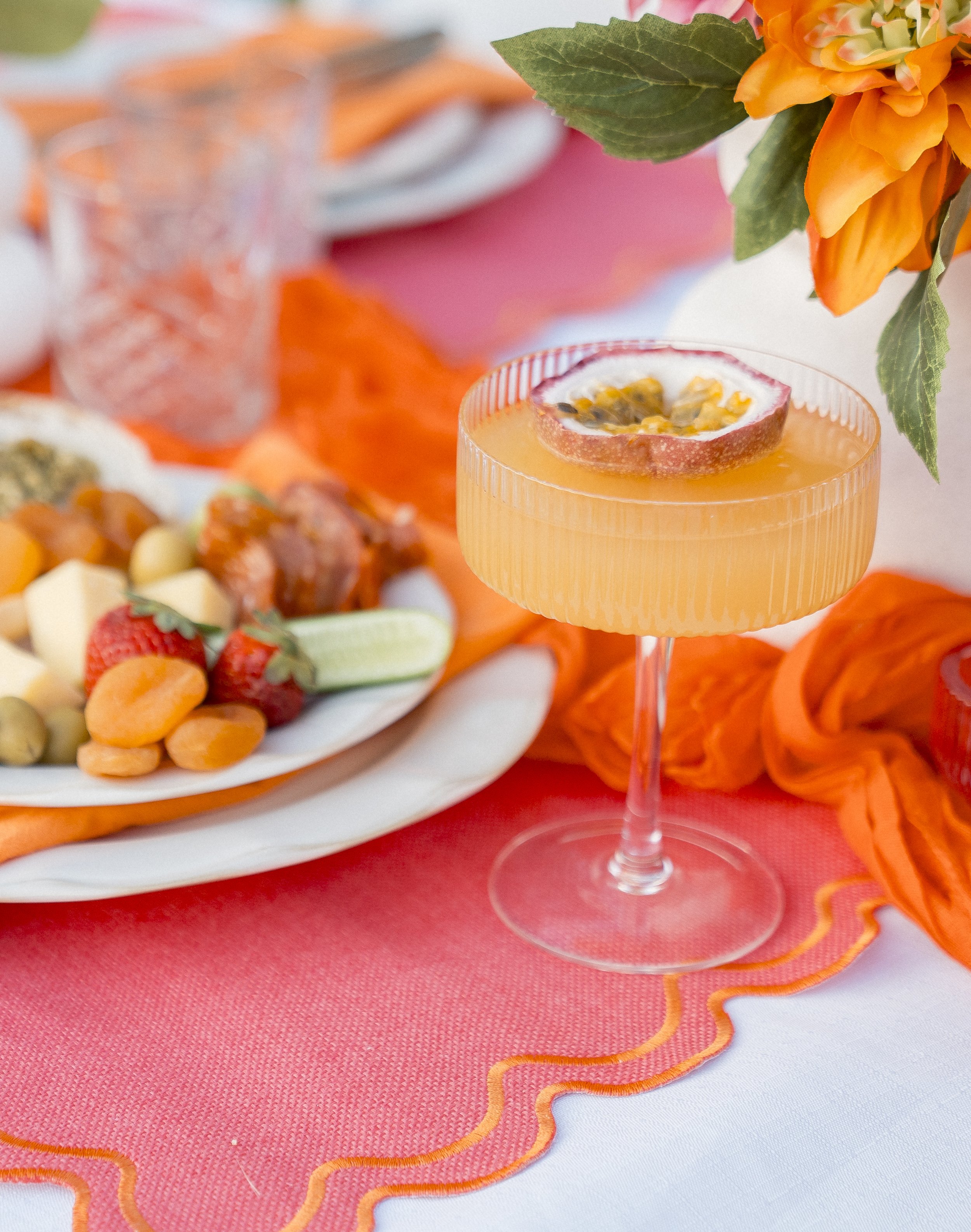 A glass of orange-colored drink garnished with a slice of passion fruit, placed on a white table with a pink napkin underneath. In the background, there is a plate of assorted fruit including strawberries, grapes, and melon, with orange and pink tabl