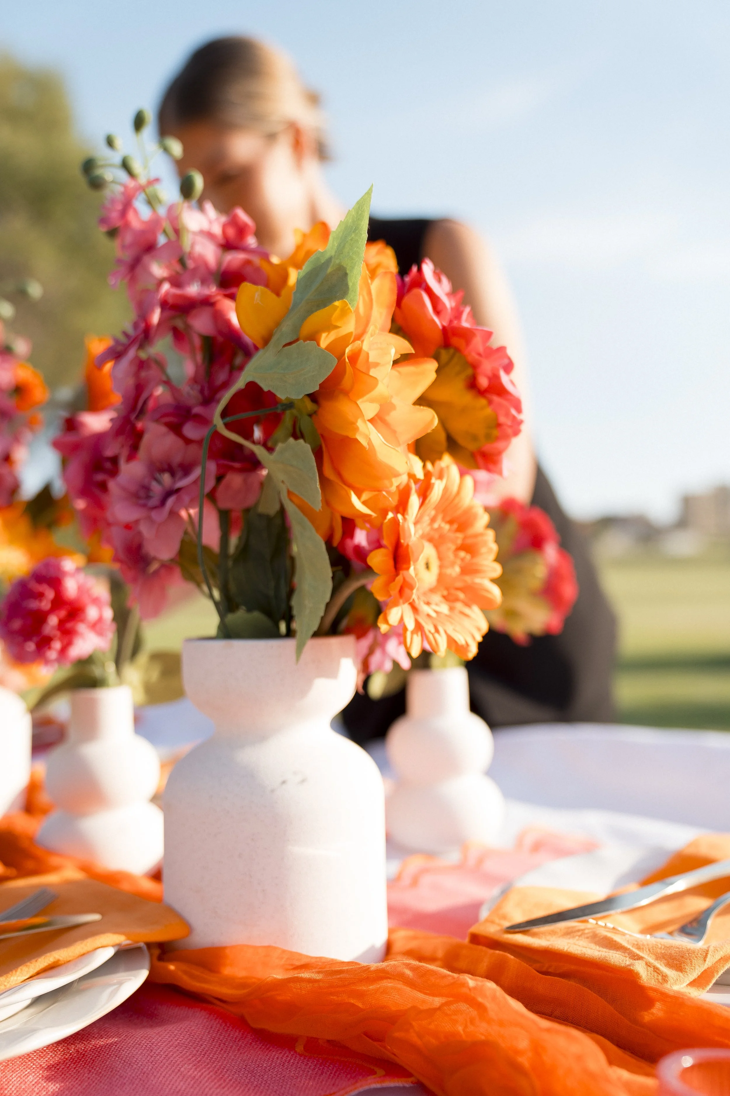 A floral centerpiece of pink, orange, and yellow flowers in white vases on an outdoor table with orange tablecloth and place settings.