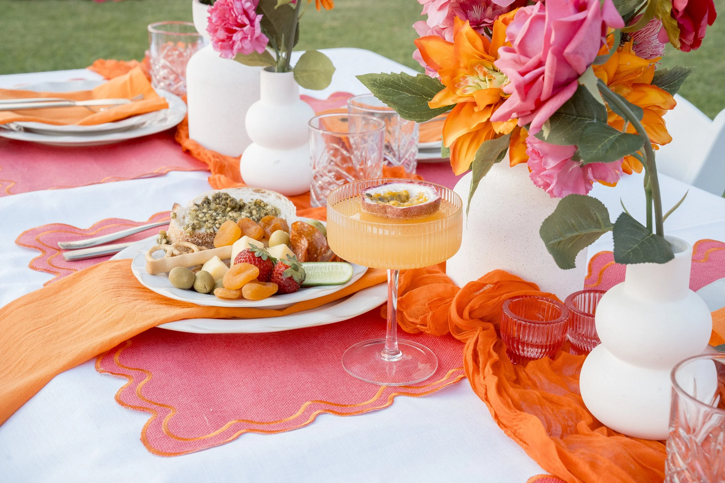 Close-up of a colorful outdoor table setting with orange, pink, and white flowers in vases, a plate of food, and glasses on a white tablecloth with orange accents.
