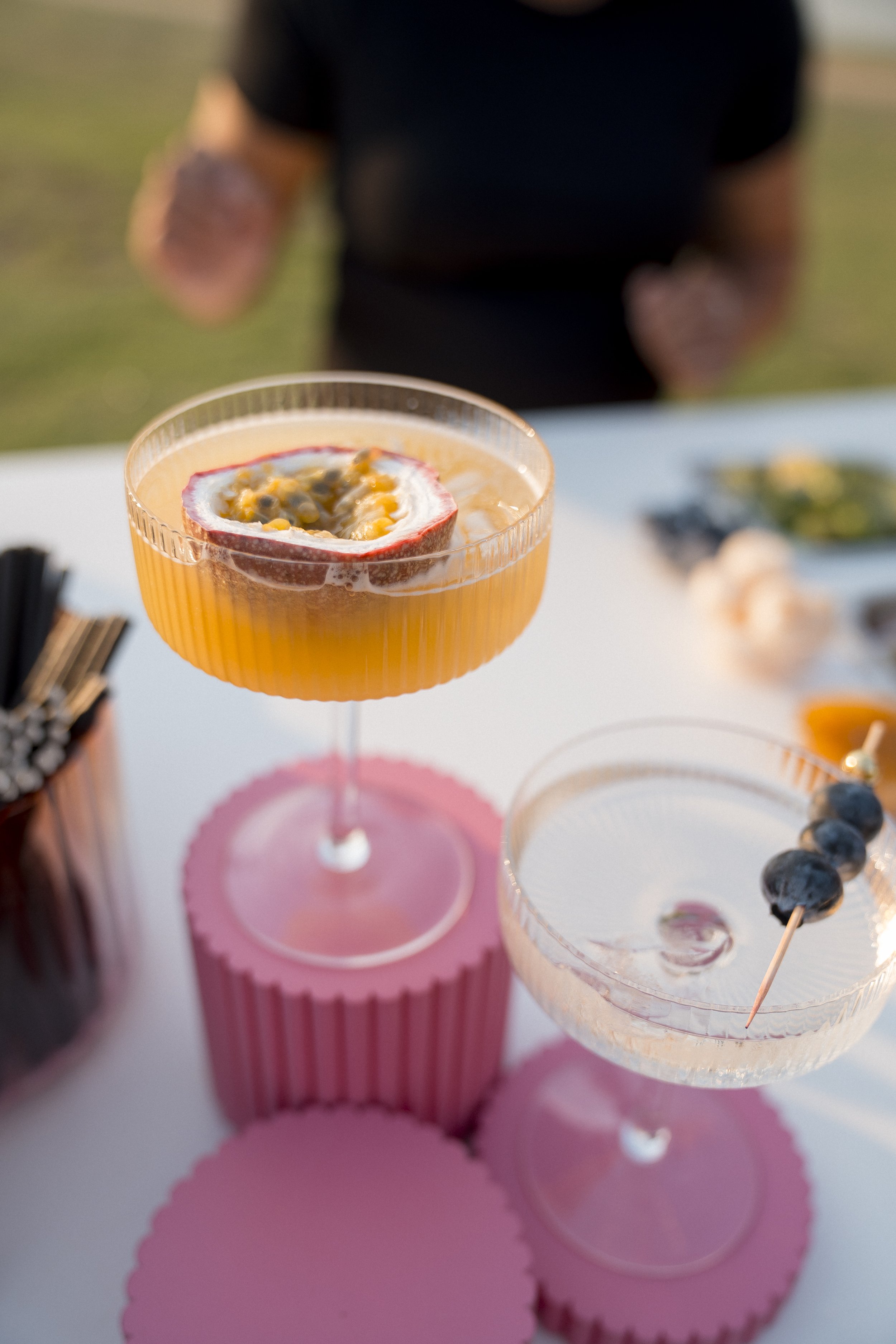 A pink cocktail glass with a passion fruit garnish, placed on a pink coaster, with other glasses and snacks in the background during a social gathering.