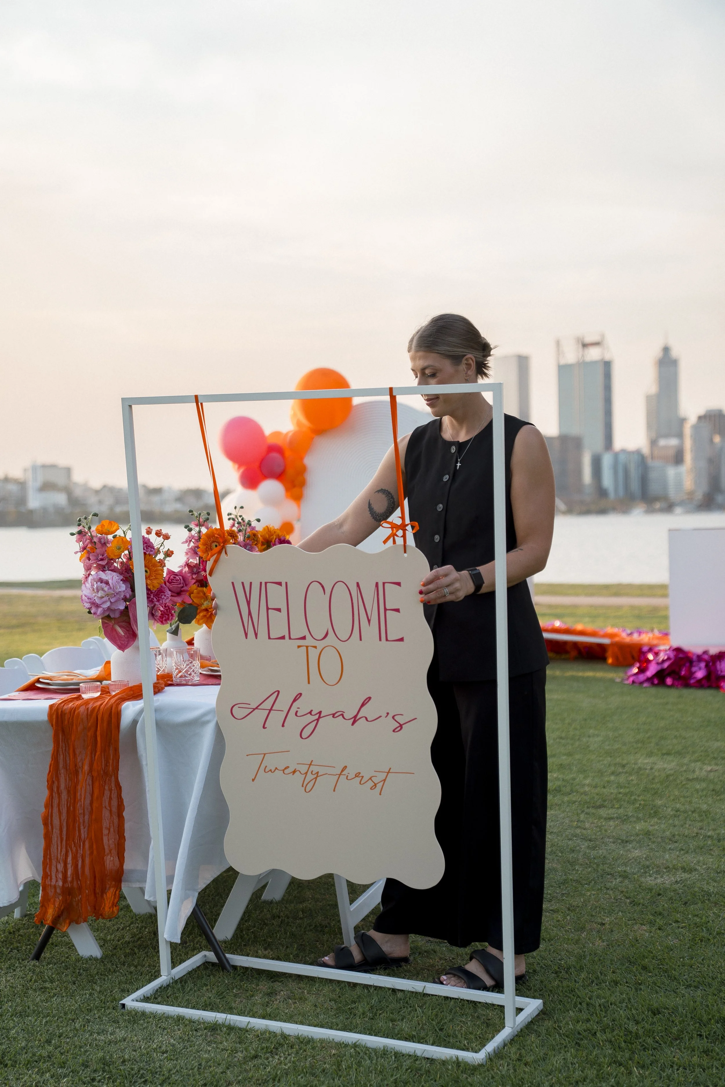Woman in black dress standing at a decorated outdoor table with a welcome sign for a twenty-fifth celebration, possibly a birthday or anniversary, on a lawn with city skyline in background during early evening.