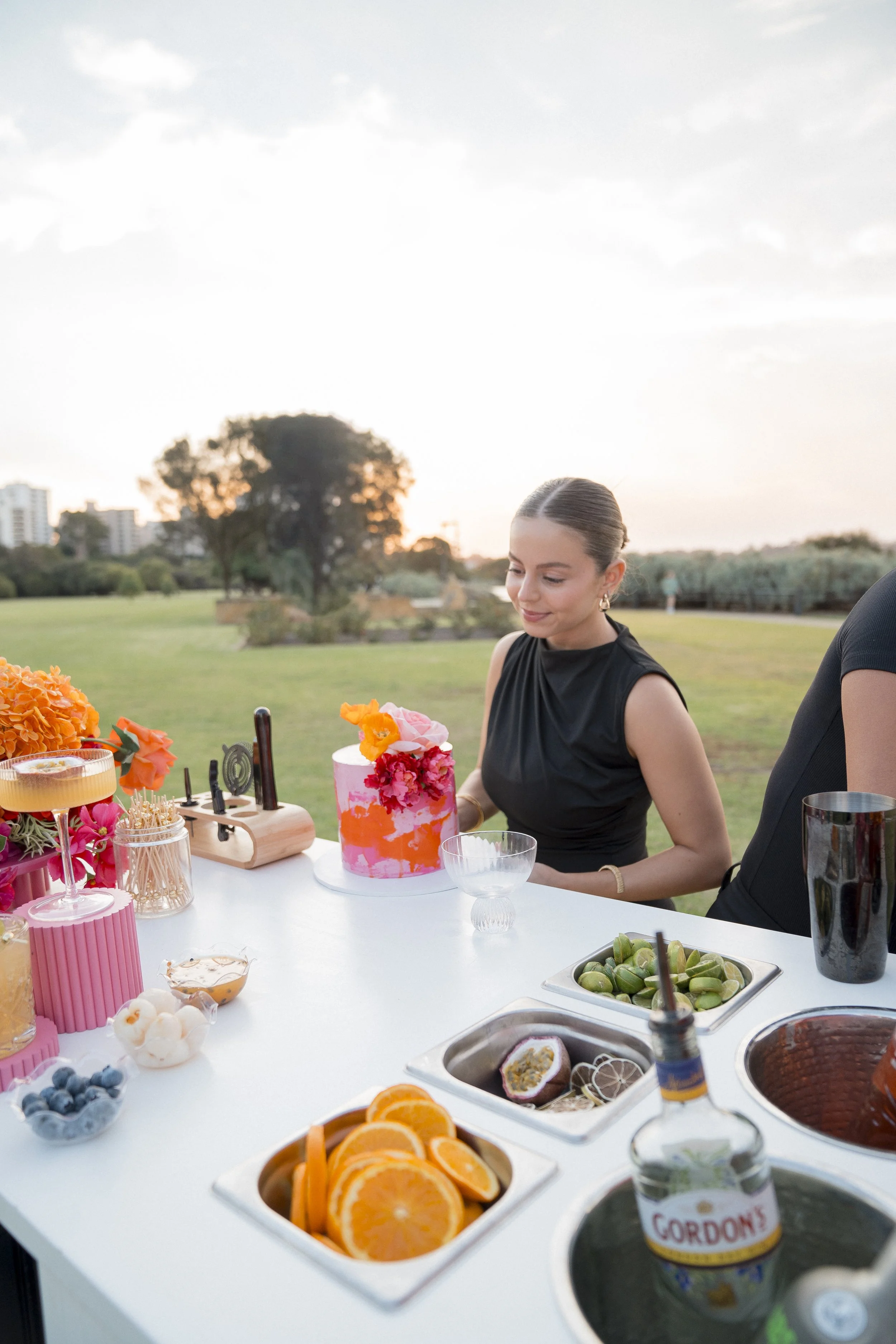A young woman in a black sleeveless dress smiling at a table with a colorful cake, fresh fruit, and drinks outdoors during sunset.