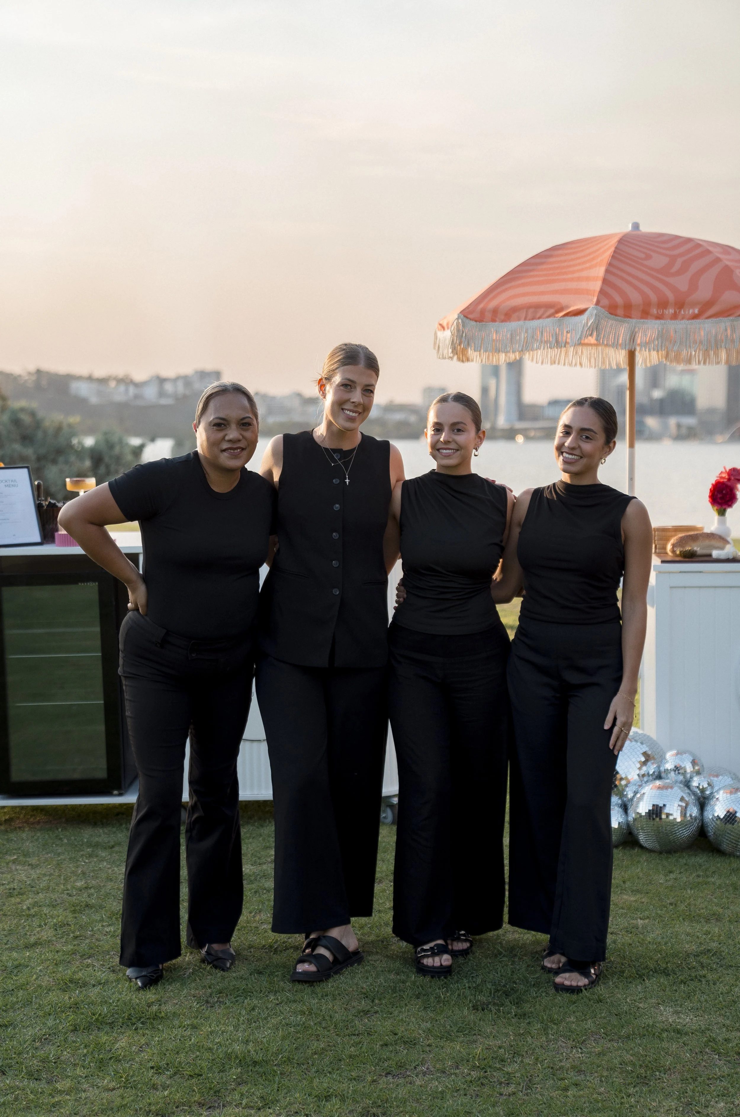 Four women standing together outdoors in front of a body of water with a cityscape in the background at sunset, dressed in black outfits.