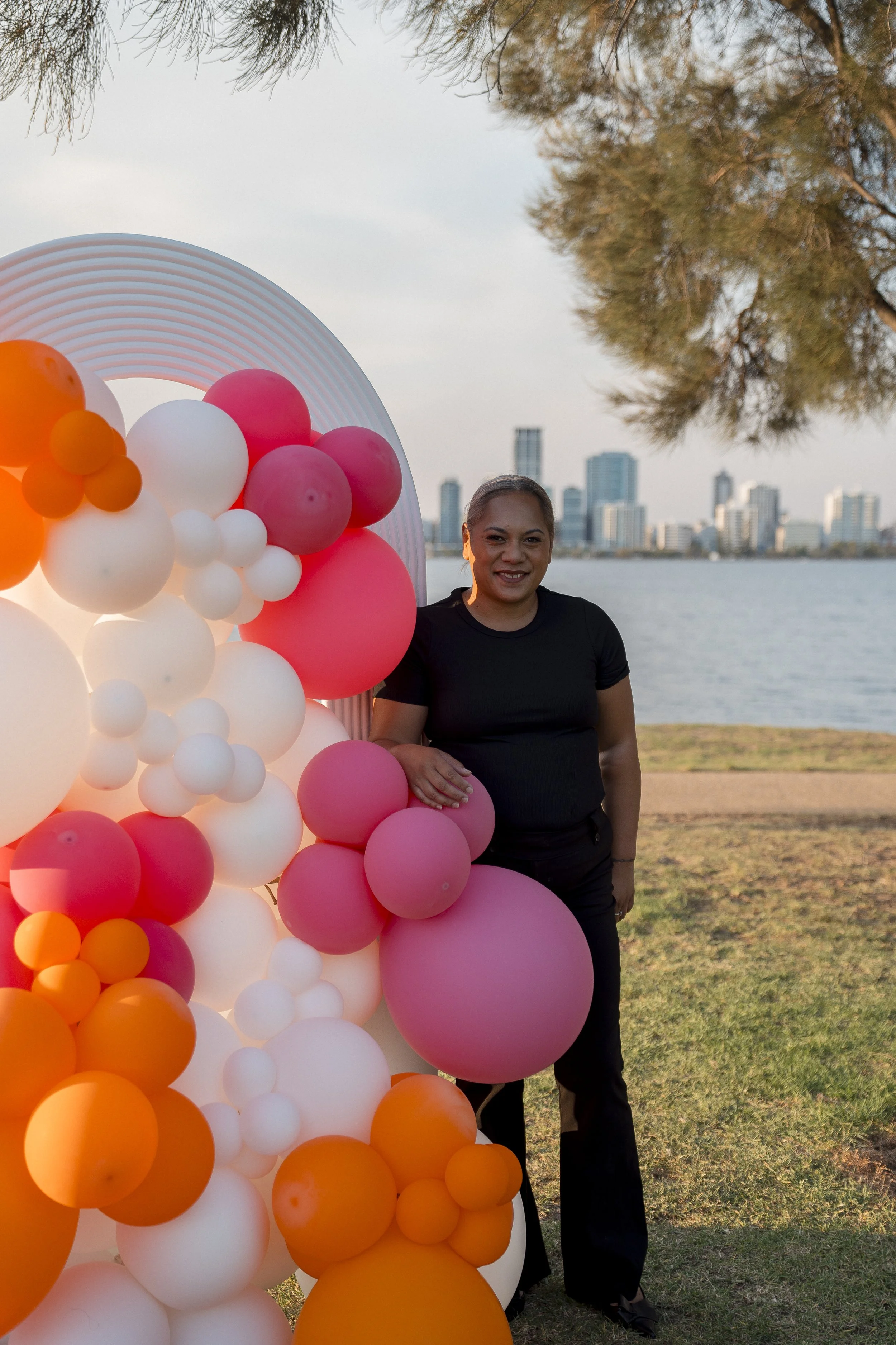 A woman in black standing near colorful balloons with city skyline and water in the background.