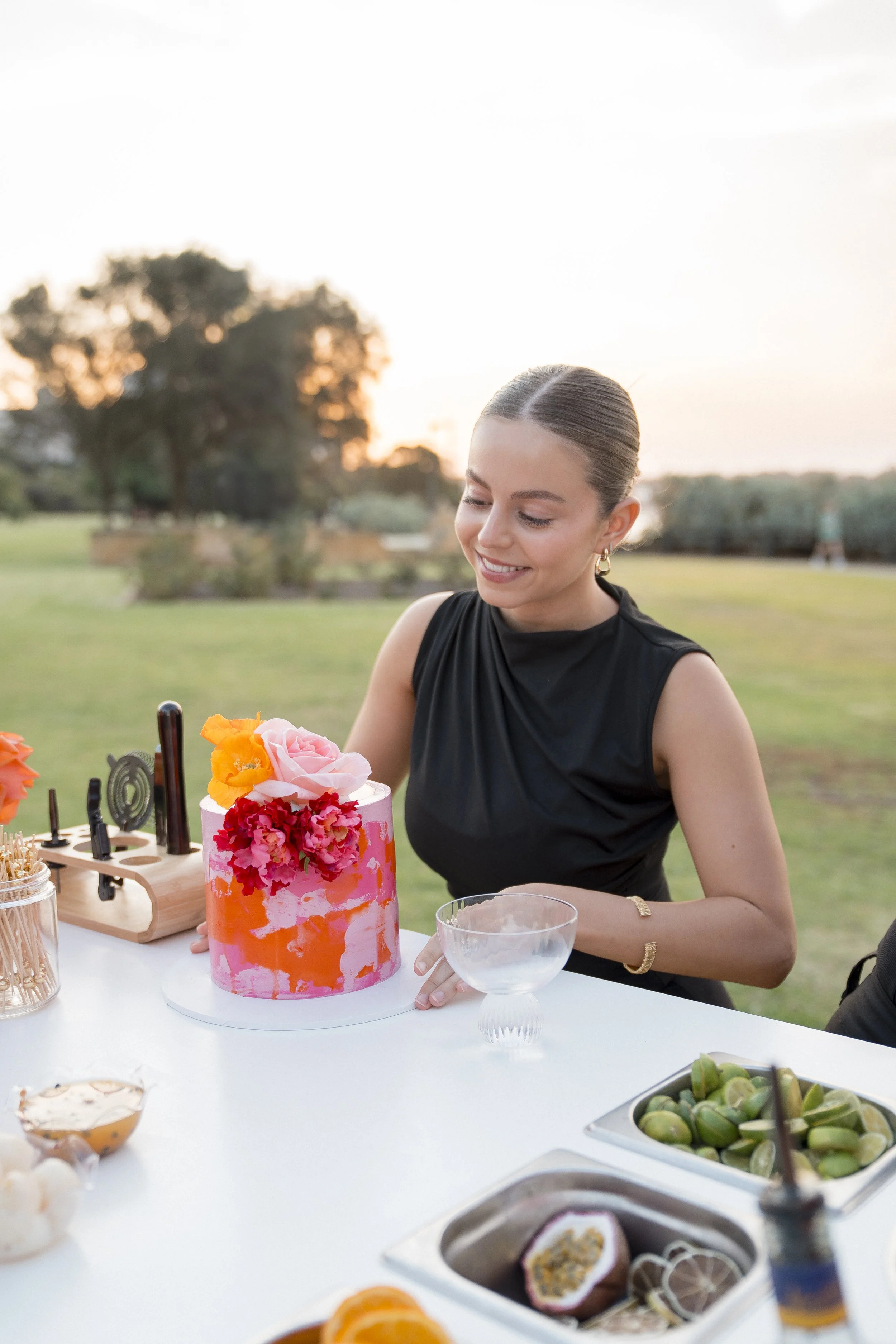 A young woman with dark hair in a bun, wearing a black sleeveless dress, is smiling at a table outdoors at sunset, surrounded by a variety of colorful foods including a pink floral cake, sushi, and fruit.
