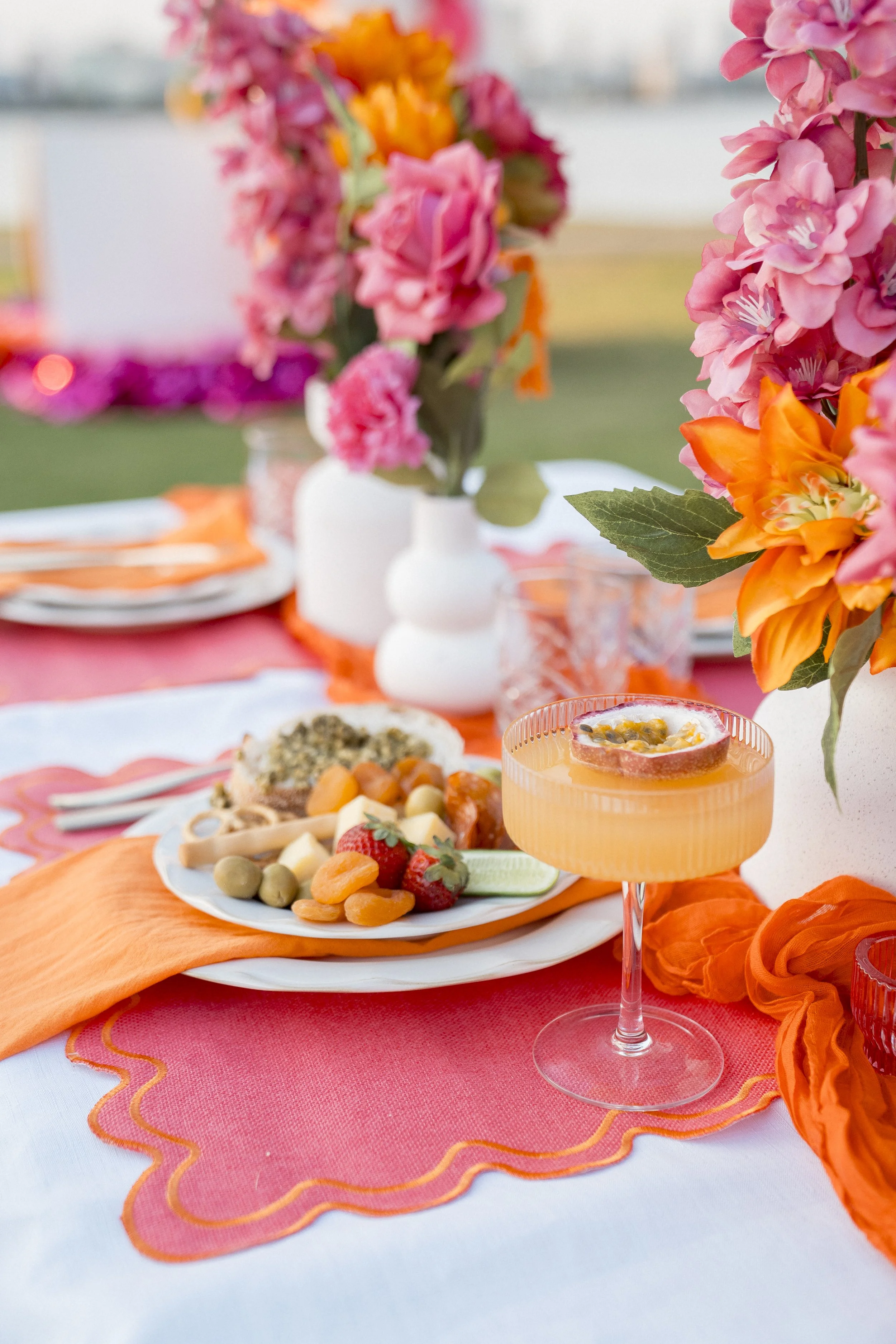 Close-up of a colorful table setting with vibrant pink, orange, and green floral arrangements, placed on an orange table runner over a pink and white tablecloth, with assorted food and drink items, including fruit, cheese, and a decorated drink glass