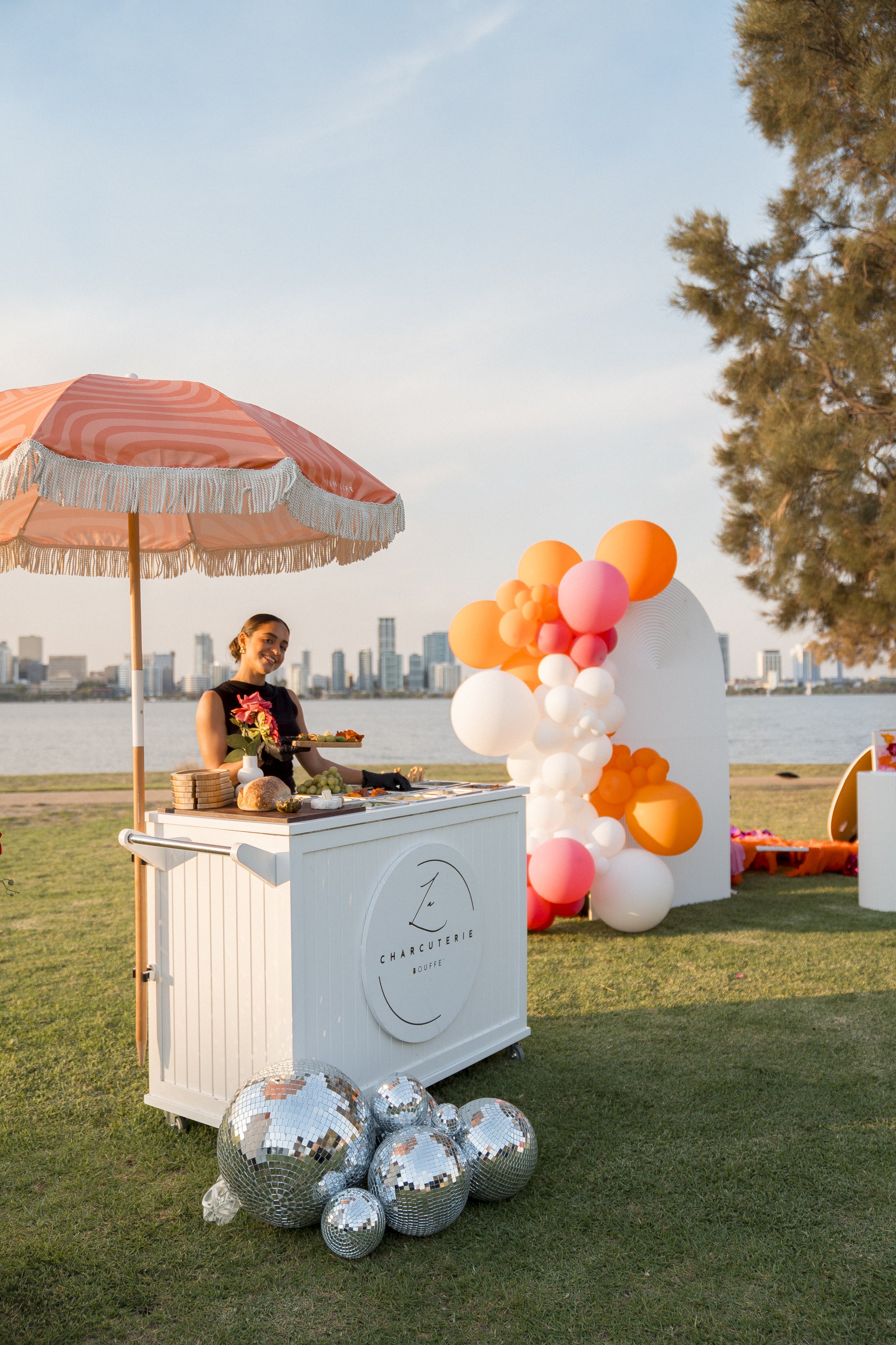 A woman standing behind a white charcuterie and cheese stand with a pink and white umbrella overhead, decorated with pink, white, and orange balloons. There are multiple disco balls on the grass in front of the stand, and a city skyline in the backgr