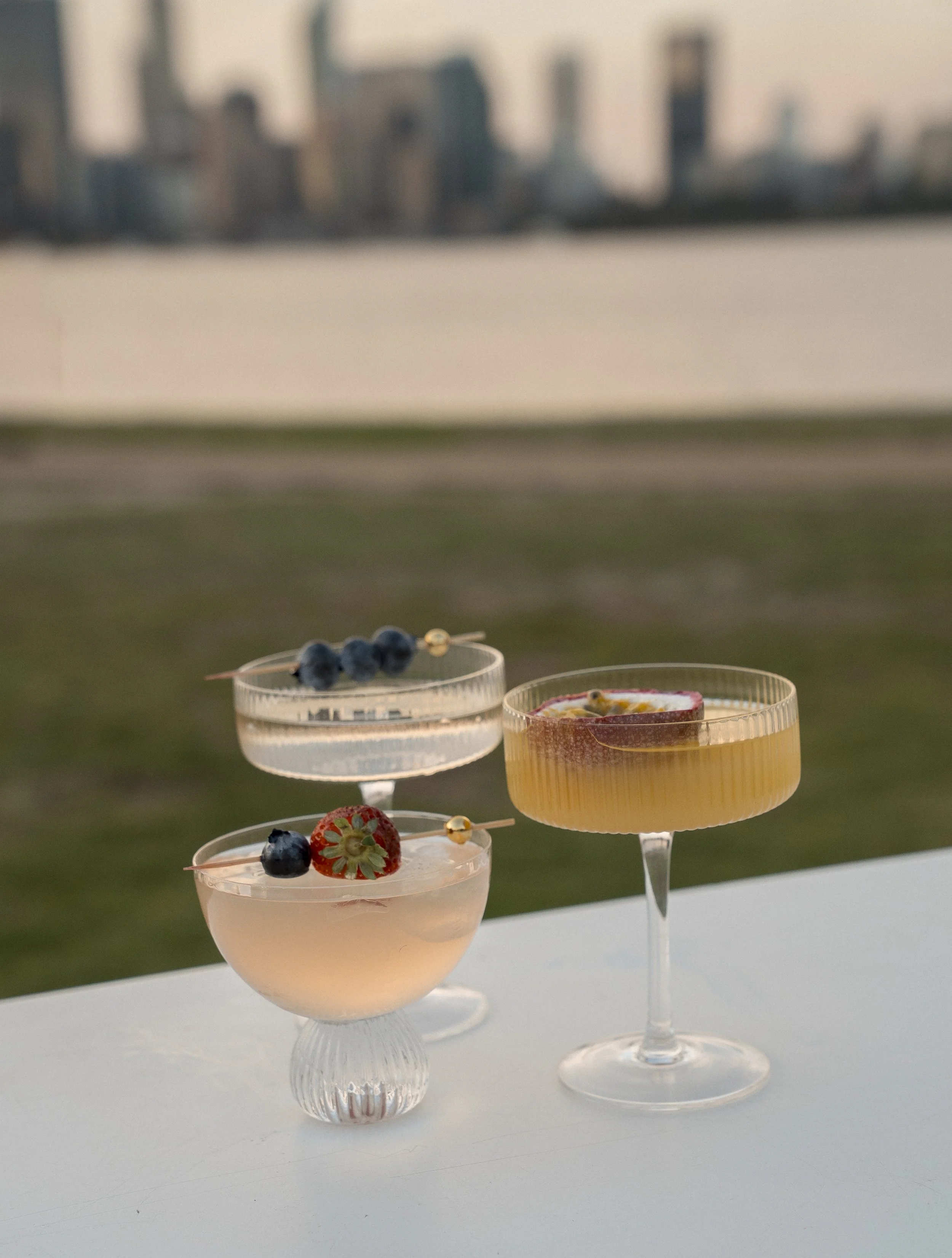 Three elegant cocktail glasses filled with pink and yellow drinks, garnished with fresh berries and fruit, set against an outdoor cityscape background at sunset.