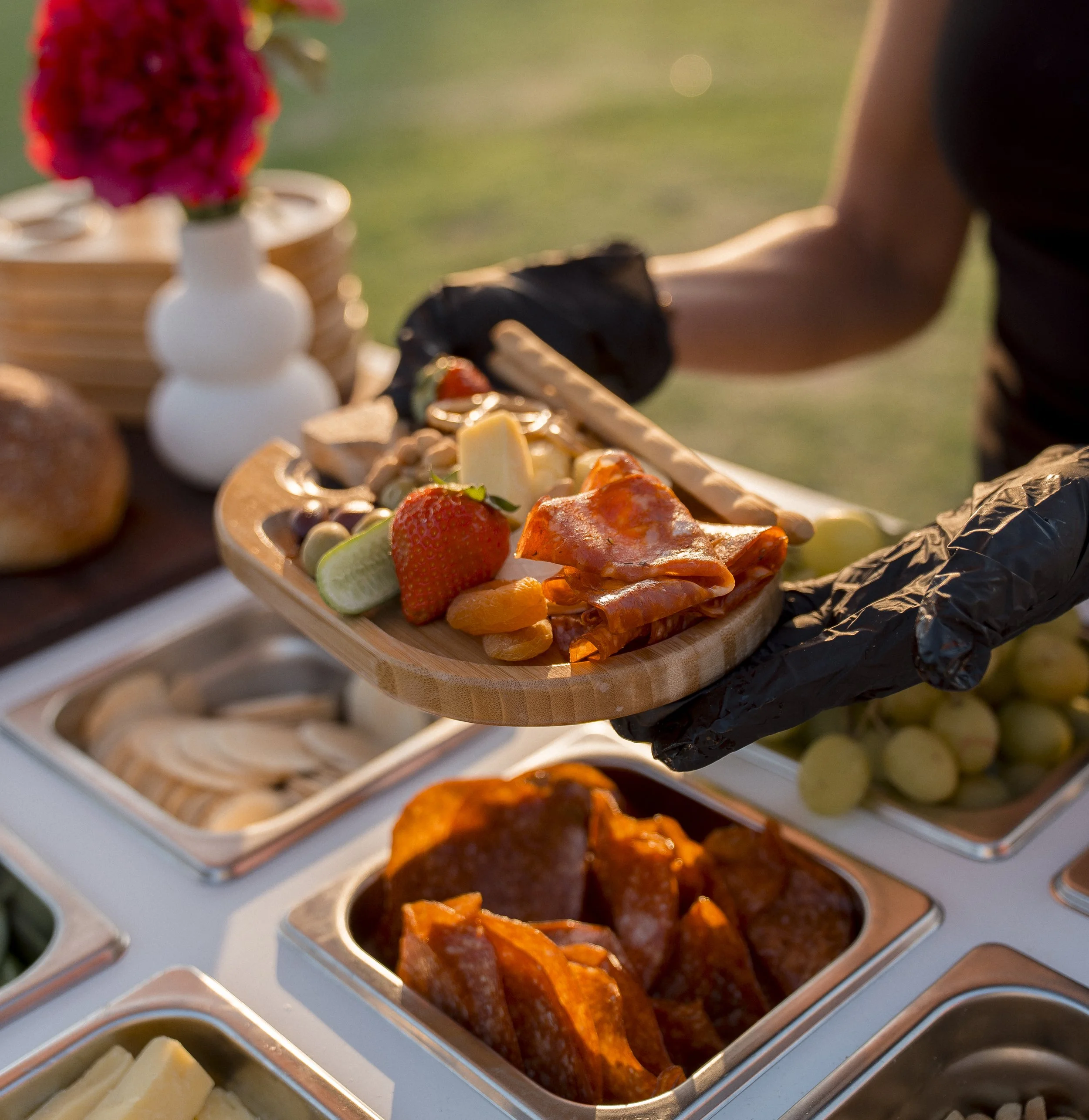 Buffet style food station with various dishes including sliced meats, cheese, strawberries, and vegetables, set outdoors with a blurred background.