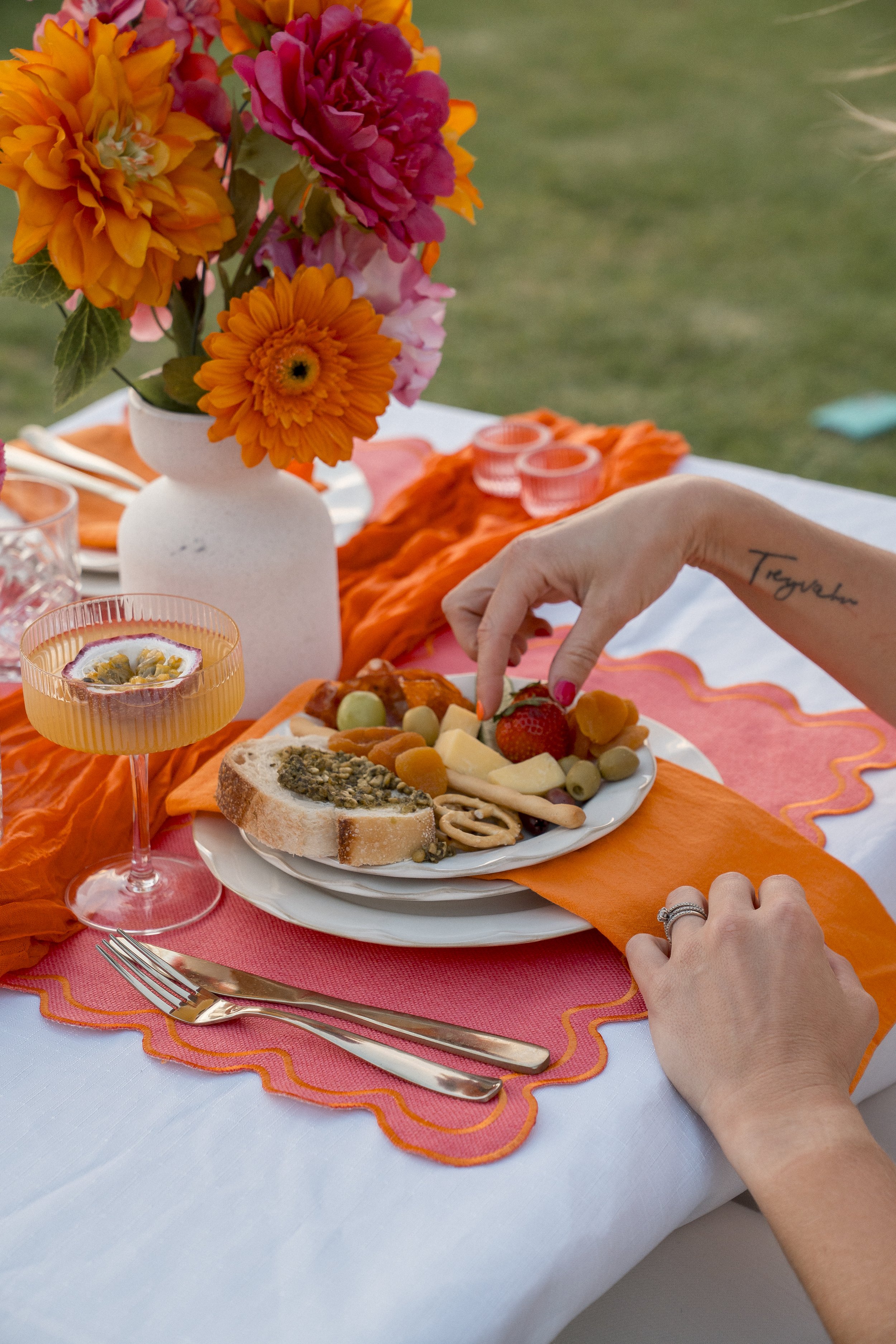 A table set with a bouquet of colorful flowers in a white vase, a plate of assorted cheese, grapes, strawberries, and crackers, pink and orange glassware, and a pink and orange tablecloth with a white tablecloth underneath.