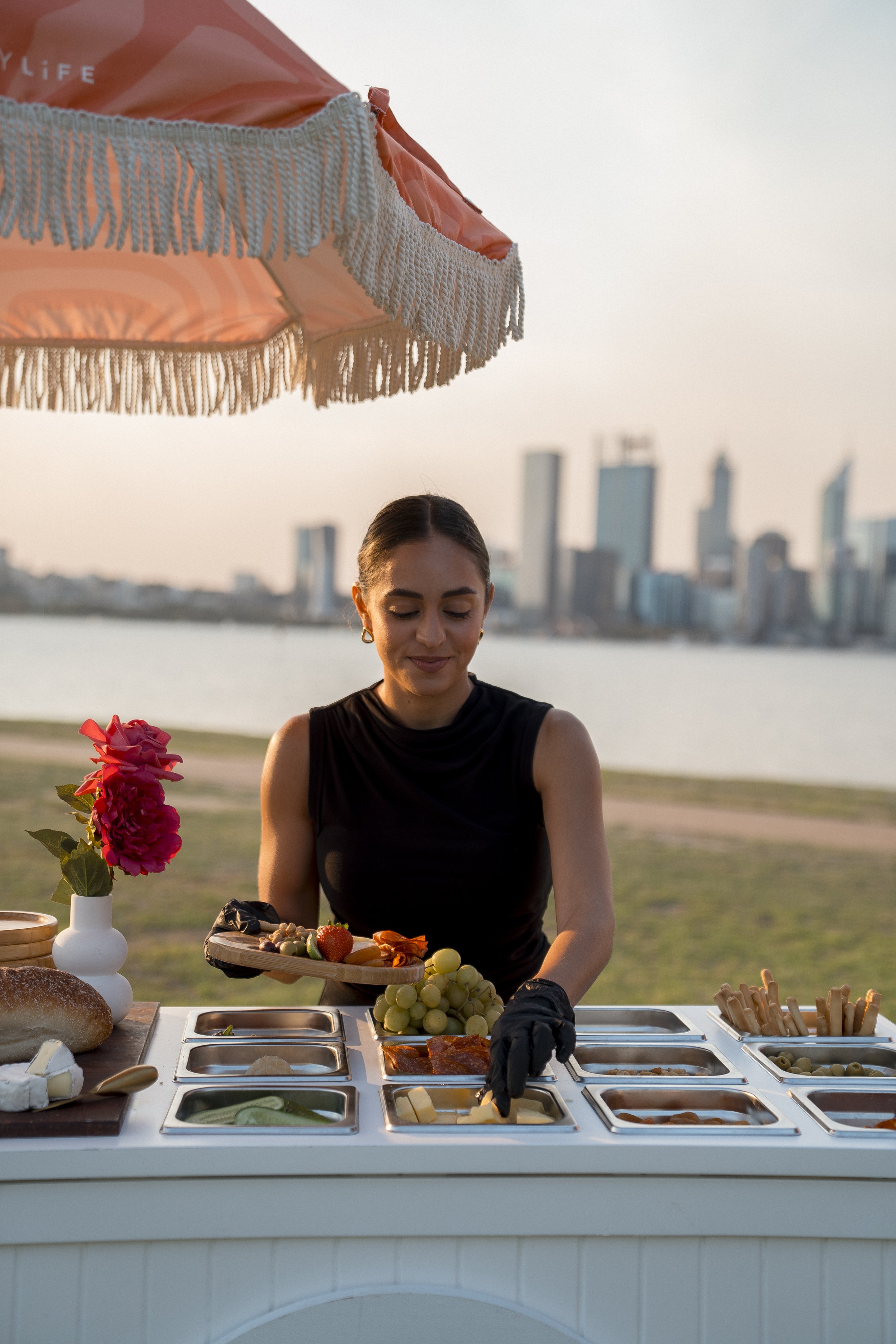 Woman wearing a black dress preparing food at an outdoor buffet table during sunset, with a city skyline in the background and a pink parasol overhead.